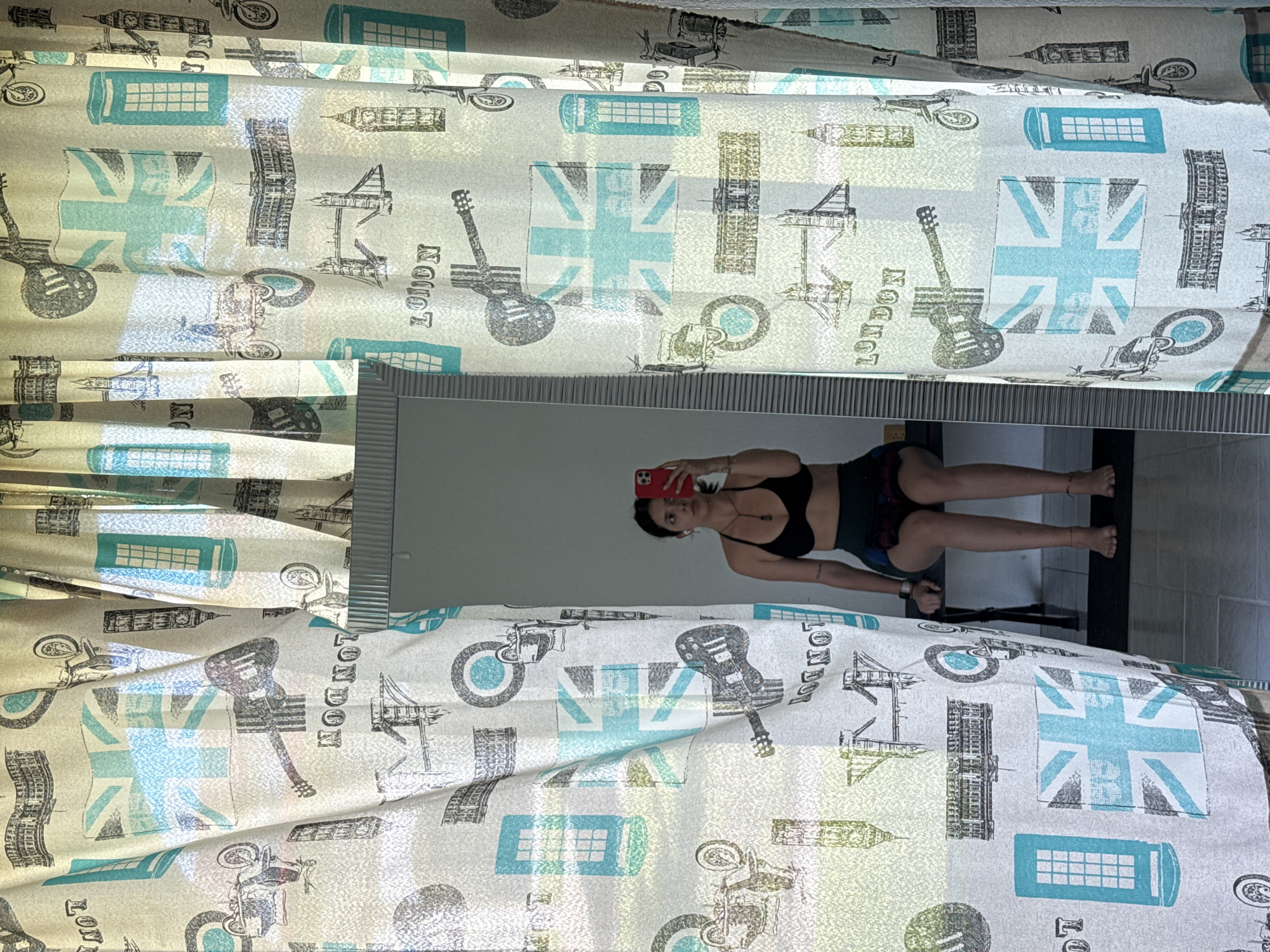 A vertical, medium-shot photograph displays a woman reflected in a mirror, flanked by curtains with a London-themed print, against a tile wall, within an interior space. The curtains, displaying Union Jack flags, London landmarks (Big Ben, Tower Bridge), London phone boxes, motorcycles, guitars, and the word "LONDON," drape on either side of the mirror. The woman in the reflection holds a red cellphone, presumably taking the photograph. She is wearing a black sports bra and patterned shorts. Behind her in the reflection is a dark object, possibly a chair, and a tile wall. The location and city cannot be determined solely from the image.