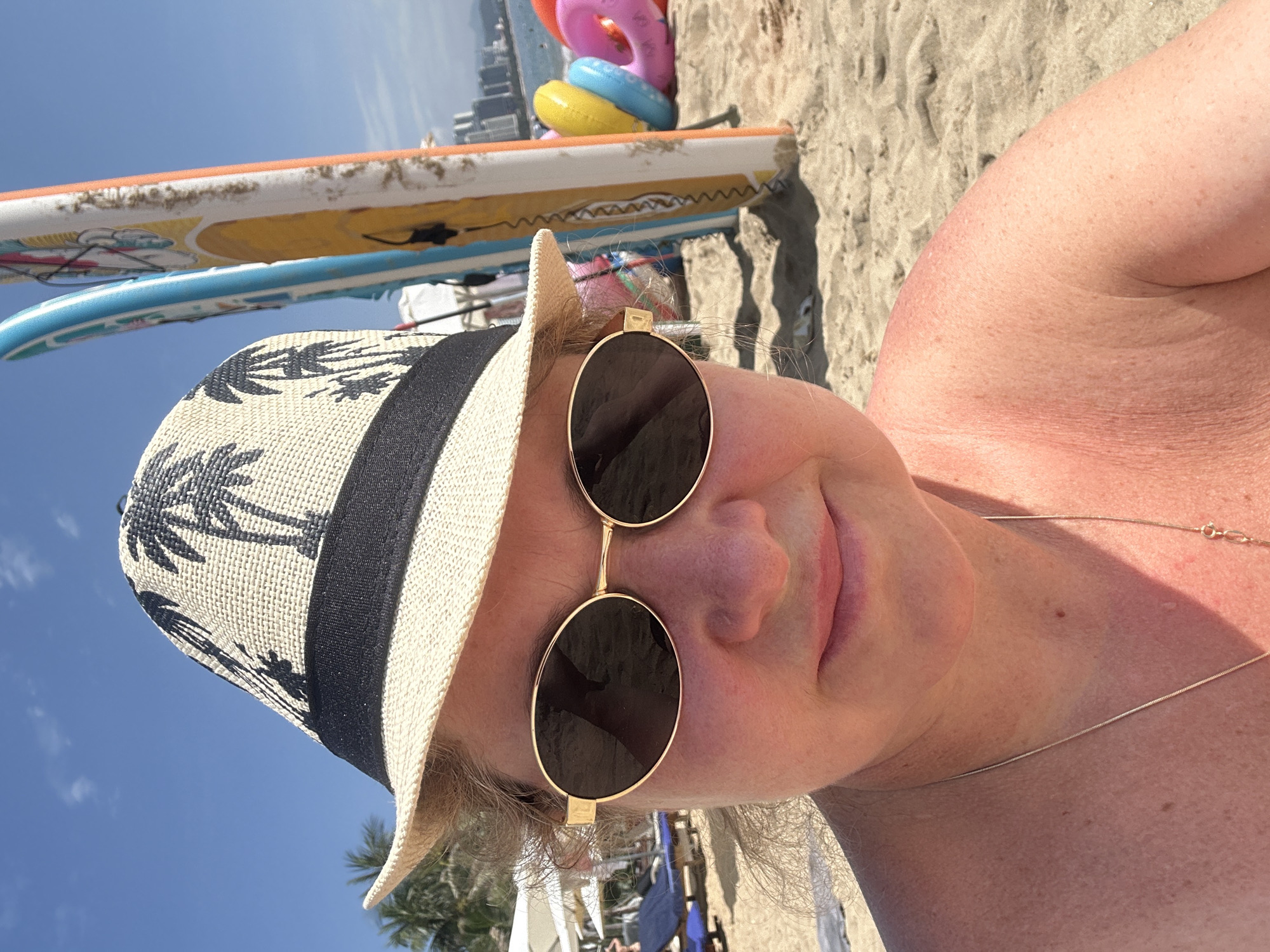 A close-up, rotated selfie shows a person with fair, sun-kissed skin at a beach in Nha Trang Ward, Vietnam, on a bright, sunny day. The individual is wearing a straw hat with a black band and black palm tree patterns, along with gold-framed, round sunglasses. A thin silver chain necklace is visible on their chest.
The background reveals a sandy beach with several leisure items. Two surfboards or paddleboards, one white with yellow and blue designs and another blue with white patterns, are propped upright in the sand. Behind them, a stack of colorful inflatable rings (pink, blue, and yellow) is visible. The distant horizon features a clear blue sky with a few wispy clouds, and a faint outline of buildings, suggesting a city near the beach. The overall impression is one of relaxation in a tropical, coastal environment. No visible text is present.