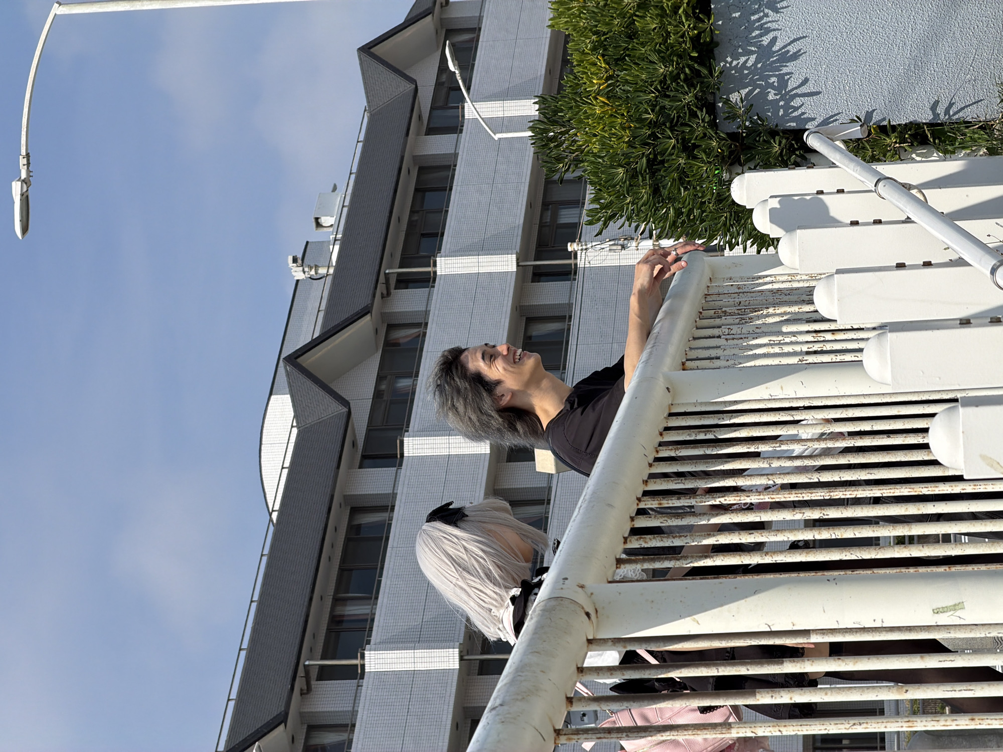 Two individuals, one with short, light-colored hair and a black outfit, and another with dark, styled hair and a black t-shirt, stand behind a white metal railing. The individual with light hair is partially obscured, facing away from the camera, while the person in the black t-shirt is looking to the left and smiling. They are positioned in front of a white building with dark, gabled roofs, which appears to be an apartment complex. A street lamp with a white fixture arcs overhead. Lush green bushes are visible to the right of the railing. This scene is located in Fujisawa, Japan, specifically near the Katase-Kaigan Station, characterized by its coastal architecture and proximity to the Enoshima Electric Railway.