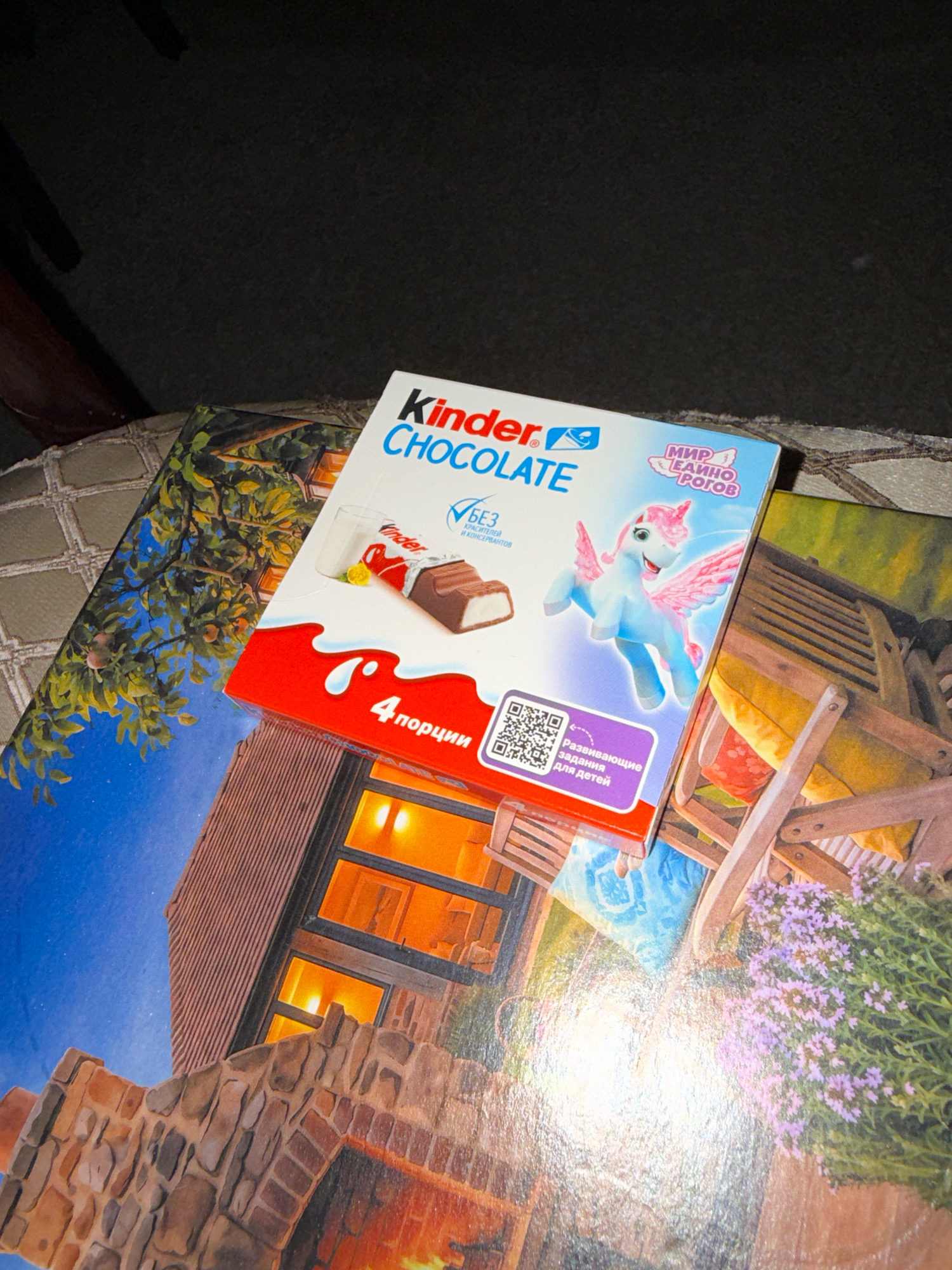 The image shows a Kinder Chocolate box placed on top of another image that appears to be a printed advertisement or a piece of artwork. The Kinder Chocolate box is red and white and features a depiction of a chocolate bar, a glass of milk, and a winged unicorn. Text on the box includes "Kinder CHOCOLATE," "4 порции" (4 portions), and a QR code with Russian text that translates to "Developing tasks for children." The background image beneath the Kinder box shows a rustic scene with a stone fireplace, a wooden structure, and flowering plants, suggesting an outdoor or cozy interior setting. The lighting is dim, and the overall scene is likely indoors. The image does not provide any specific information that would allow for its location to be determined in Zavolzhye, Russia.