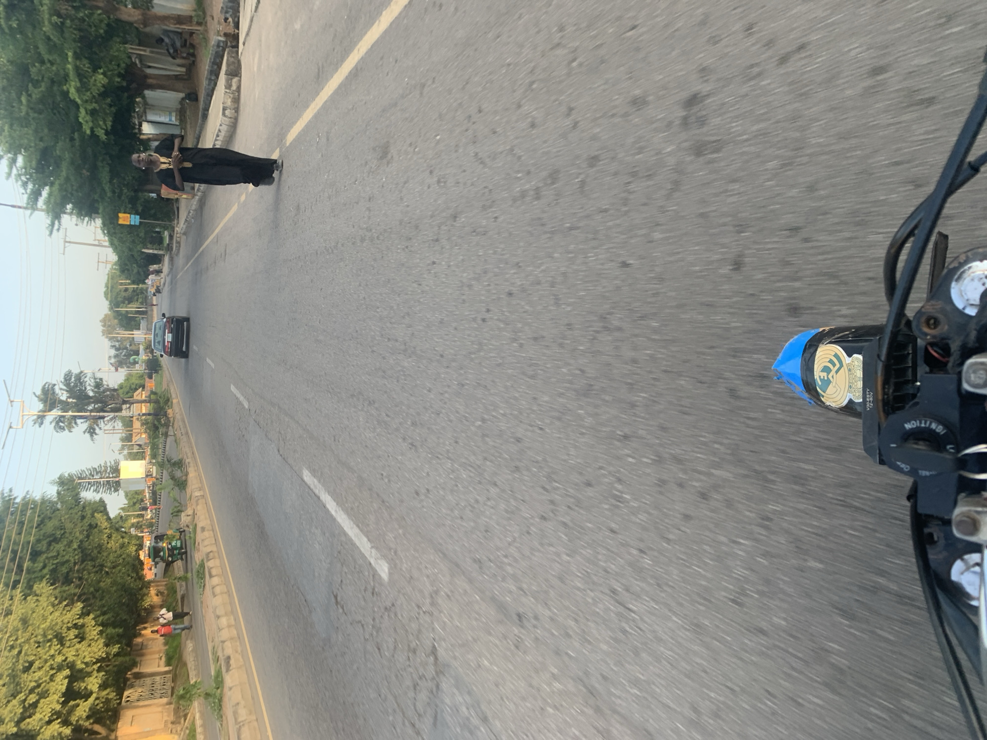 This is a first-person perspective, likely from a motorcycle or tricycle, capturing a street scene in Jos, Nigeria. The road is paved with asphalt and marked with a white dashed line on the left and a solid yellow line on the right. The environment appears to be a moderately busy urban area with buildings and vegetation visible along the sides of the road.
In the mid-ground, a person is walking in the middle of the road, wearing a long, dark robe with ornate gold embroidery on the front. They are facing towards the camera, with their hands clasped in front. Further down the road, a dark-colored sedan is traveling away from the camera. On the left side of the road, two individuals are walking on the sidewalk, and a green auto-rickshaw (Keke Napep) is parked.
The foreground is dominated by the handlebars and instrument panel of a motorcycle or similar vehicle, with a blue cover visible on the seat. The image has a slight motion blur, indicating movement. The lighting suggests it is either early morning or late afternoon.