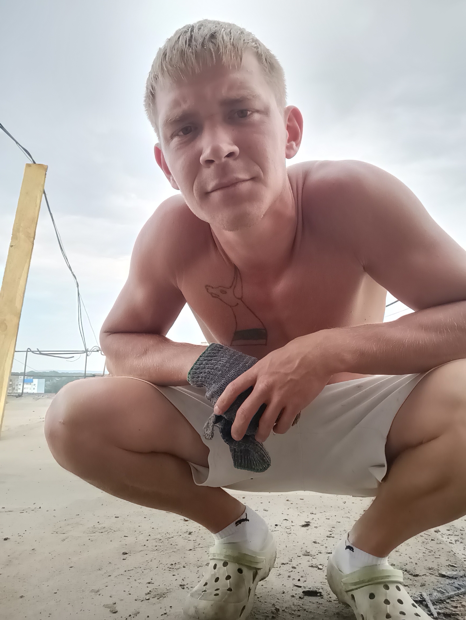 A young man with short, blond hair is crouching down, looking at the camera with a neutral expression. He is shirtless, wearing light-colored shorts, white socks, and light-colored clogs with holes. He is holding a dark-colored glove in his right hand. A tattoo is visible on his chest.
The setting appears to be outdoors, possibly on a rooftop or elevated area, with a cloudy sky overhead. There are some construction elements in the background, including a yellow pole and wires. In the distance, there are buildings, suggesting an urban or semi-urban environment.
The time of day is likely daytime, given the brightness of the sky. The weather appears overcast. The image was taken in Chaikovsky, Russia. There is no visible text in the image.