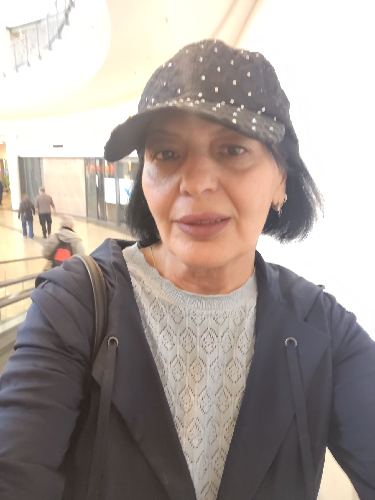 A woman is the primary subject of a selfie taken in what appears to be a shopping mall, possibly in Kehl, Germany.
The woman is wearing a dark, possibly sequined, baseball cap, a light-colored patterned sweater, and a dark blazer or jacket. She has short, dark hair and is wearing hoop earrings. She appears to be standing on an elevated platform or escalator, as people are visible walking below.
The background shows the interior of a shopping mall. There are storefronts with brightly lit interiors. A staircase or escalator can be seen in the upper left corner of the image. Other people are walking in the background. The light is bright and somewhat diffused.