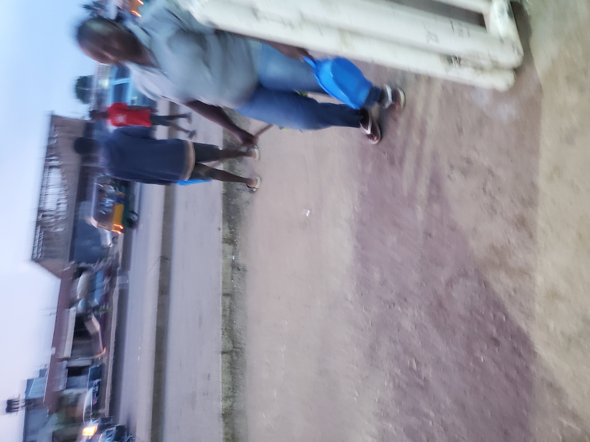 The image shows a street scene in Jos, Nigeria, with several people and vehicles visible. In the foreground, a woman wearing a grey polo shirt and blue jeans is holding a blue dustpan. To her left, two young men are standing on the side of the road. One is wearing a dark blue hooded sweatshirt and shorts, while the other is wearing a red t-shirt and shorts. Behind them, a yellow and green auto-rickshaw is parked. The background shows buildings and more vehicles, suggesting a busy urban environment. The lighting indicates it is either early morning or late evening.