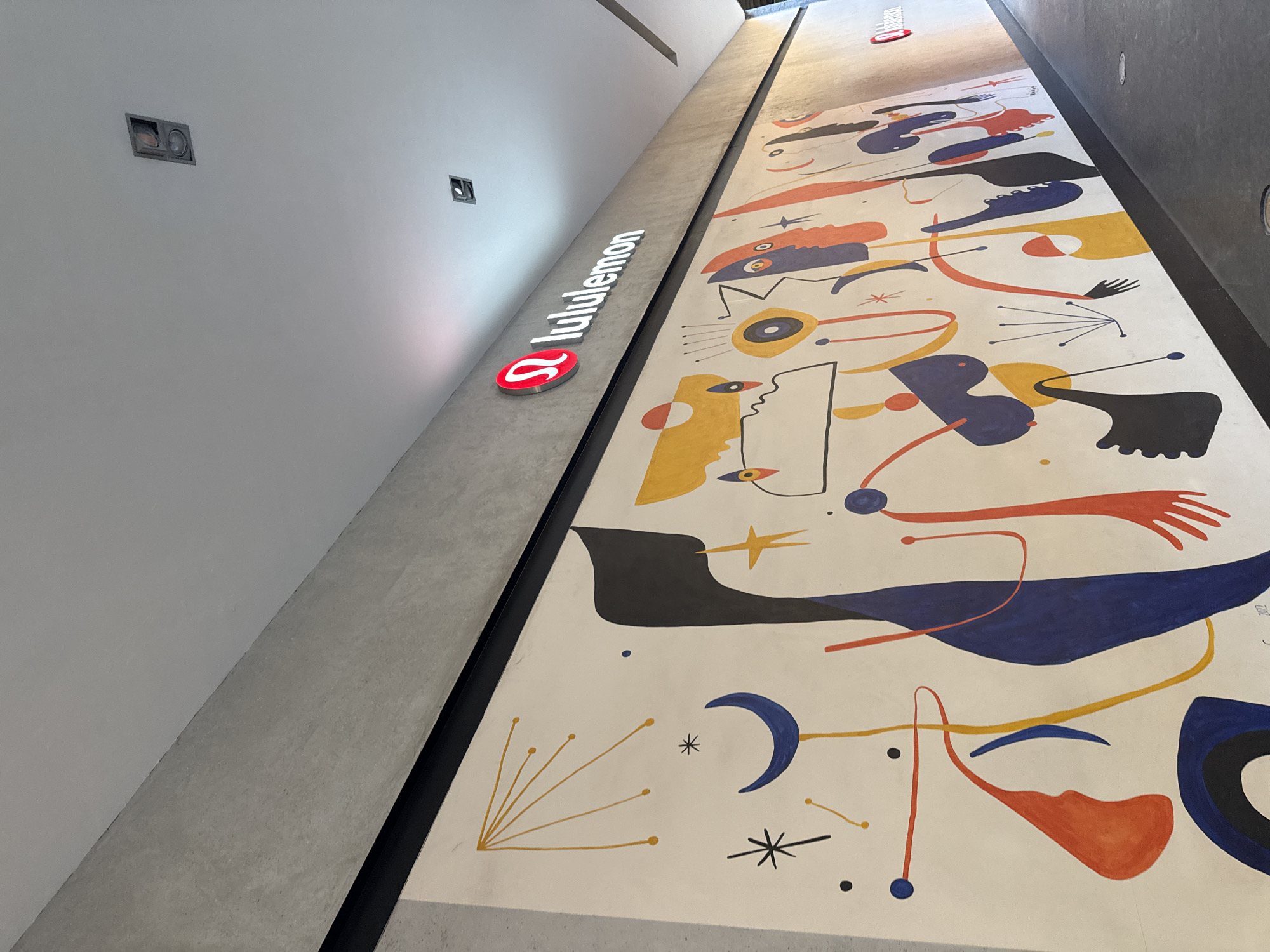 The image shows the interior of a Lululemon store in Puebla, Mexico. The wall features an abstract mural in a style reminiscent of Joan Miró, with shapes in blue, yellow, red, and black against a cream background. The "Lululemon" logo is displayed on the wall. The image is taken from a low angle.