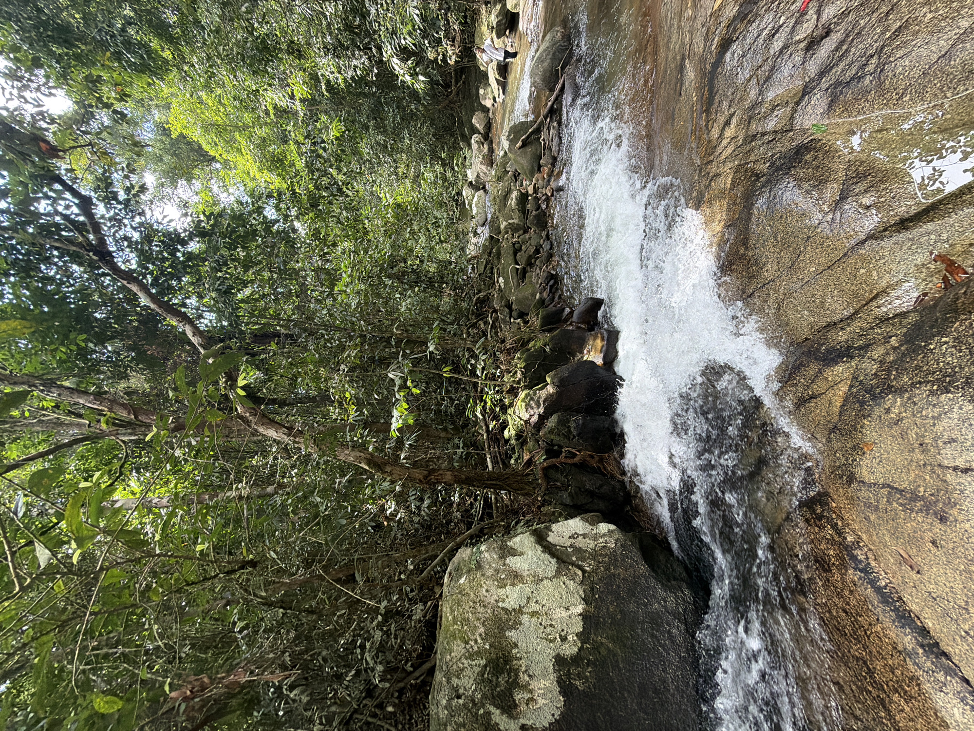 A natural scene unfolds in a jungle environment with a river flowing over rocks. The water is clear and turbulent in some areas, creating small rapids. Large rocks are scattered throughout the riverbed and along the banks, some covered in moss. Lush green trees and dense foliage form a canopy overhead, with sunlight filtering through the leaves. A person in a white shirt and dark pants stands in the river on the right side of the image, facing away from the camera, with their hand on their hip. The ground in the foreground is composed of smooth, wet rocks. The location is identified as Khuekkhak, Thailand.