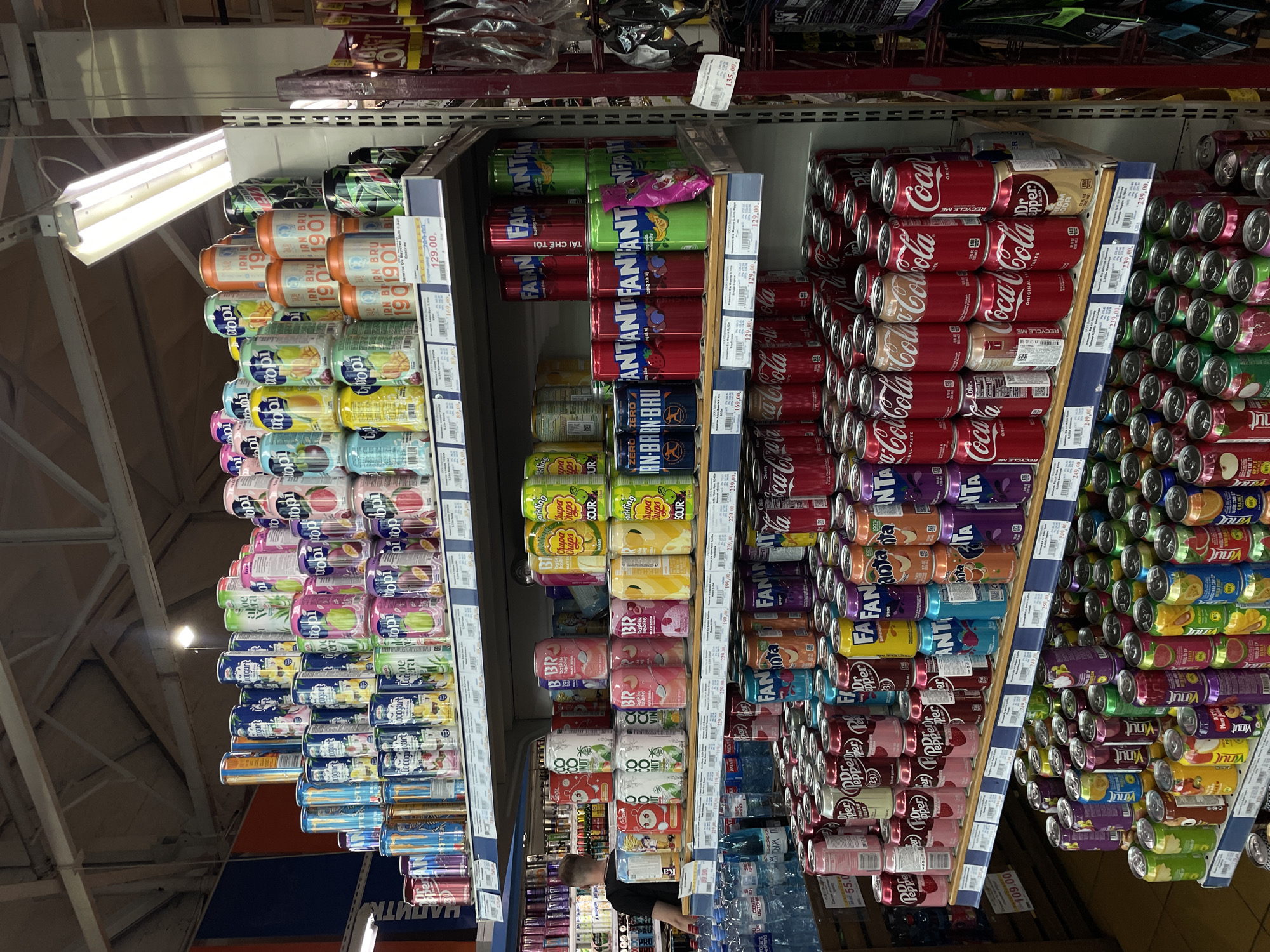This image shows a man standing in a brightly lit supermarket aisle, surrounded by shelves stocked with a wide variety of canned beverages. The shelves are organized with different brands and flavors, including Coca-Cola, Fanta, Dr. Pepper, and various fruit-flavored drinks. The lighting suggests an indoor environment, likely during daytime hours. The man appears to be looking at the products, possibly shopping or working.
Notable text visible on the cans and price tags includes brand names like "Coca-Cola," "Fanta," "Dr. Pepper," and "Vinut," as well as pricing information such as "129.00," "169.00," and "239.00." Some text also indicates "RECYCLE ME" and "ORIGINAL TASTE," providing product details. The overall scene depicts a typical retail environment with a focus on beverage products. The location context provided is Sinyavino, Russia.