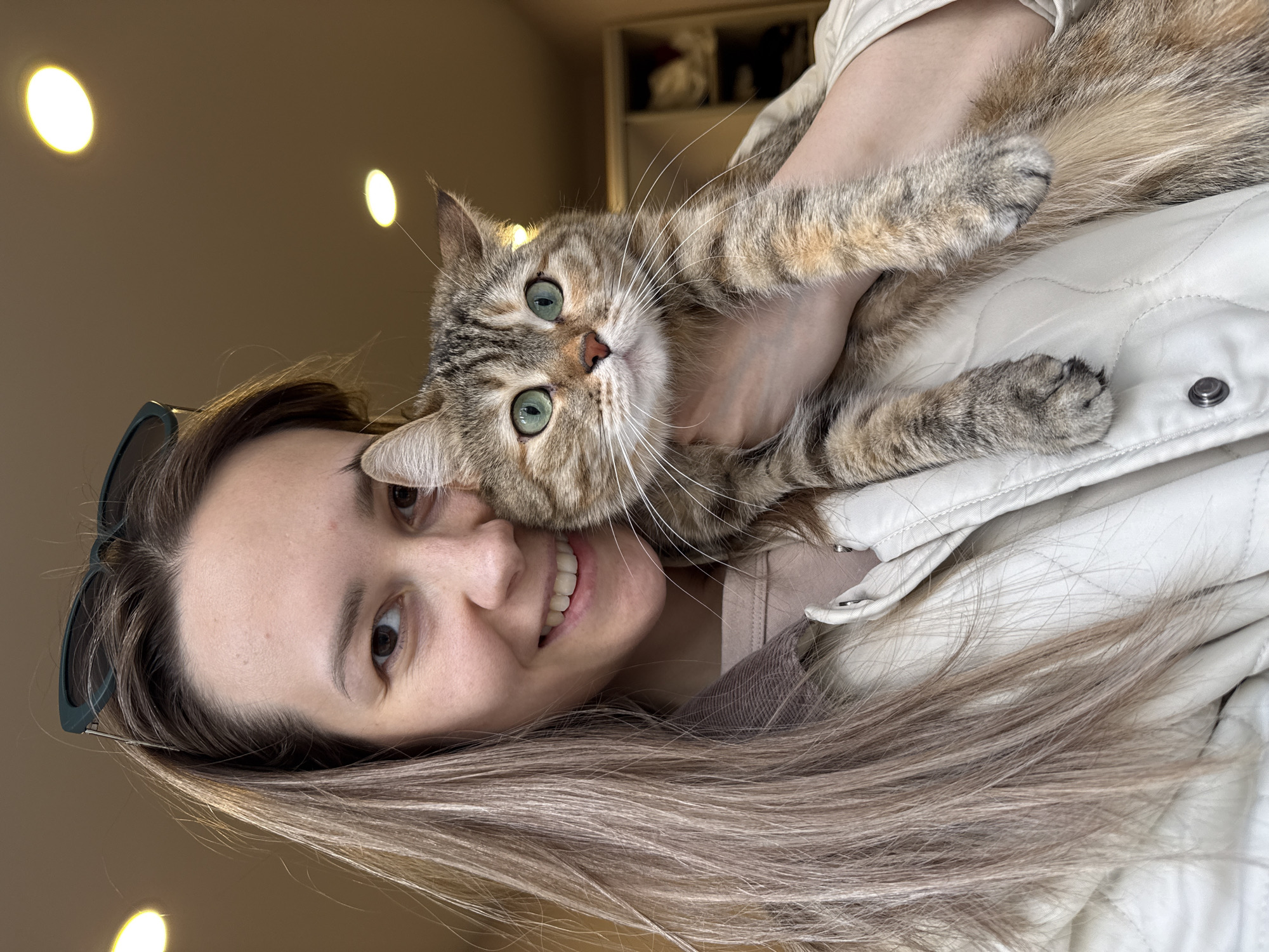 This rotated portrait captures a warm, close-up moment between a young woman and her cat indoors. The woman, positioned on the left, has long, straight brown hair with lighter highlights, dark eyes, and a broad, joyful smile revealing her teeth. She wears a light-colored, possibly off-white or beige, quilted jacket over a lighter top, with dark-rimmed sunglasses resting on her head.
Cradled in her arms, a beautiful shorthaired cat with a tabby-like pattern featuring shades of brown, grey, and warmer tones, engages directly with the camera. Its most striking feature is a pair of vivid bright green eyes. The cat also has long, prominent white whiskers and its paws are visible, gently resting on the woman's arm and jacket.
The background suggests a home environment, with a warm, neutral-toned wall. Two circular, warm-toned light fixtures are visible on the upper left wall, indicating indoor lighting. In the upper right background, indistinct shelves or cabinetry can be seen. The image conveys a sense of affection and happiness between the woman and her pet. No text is visible in the image. The scene is located in Chaikovsky, Russia.