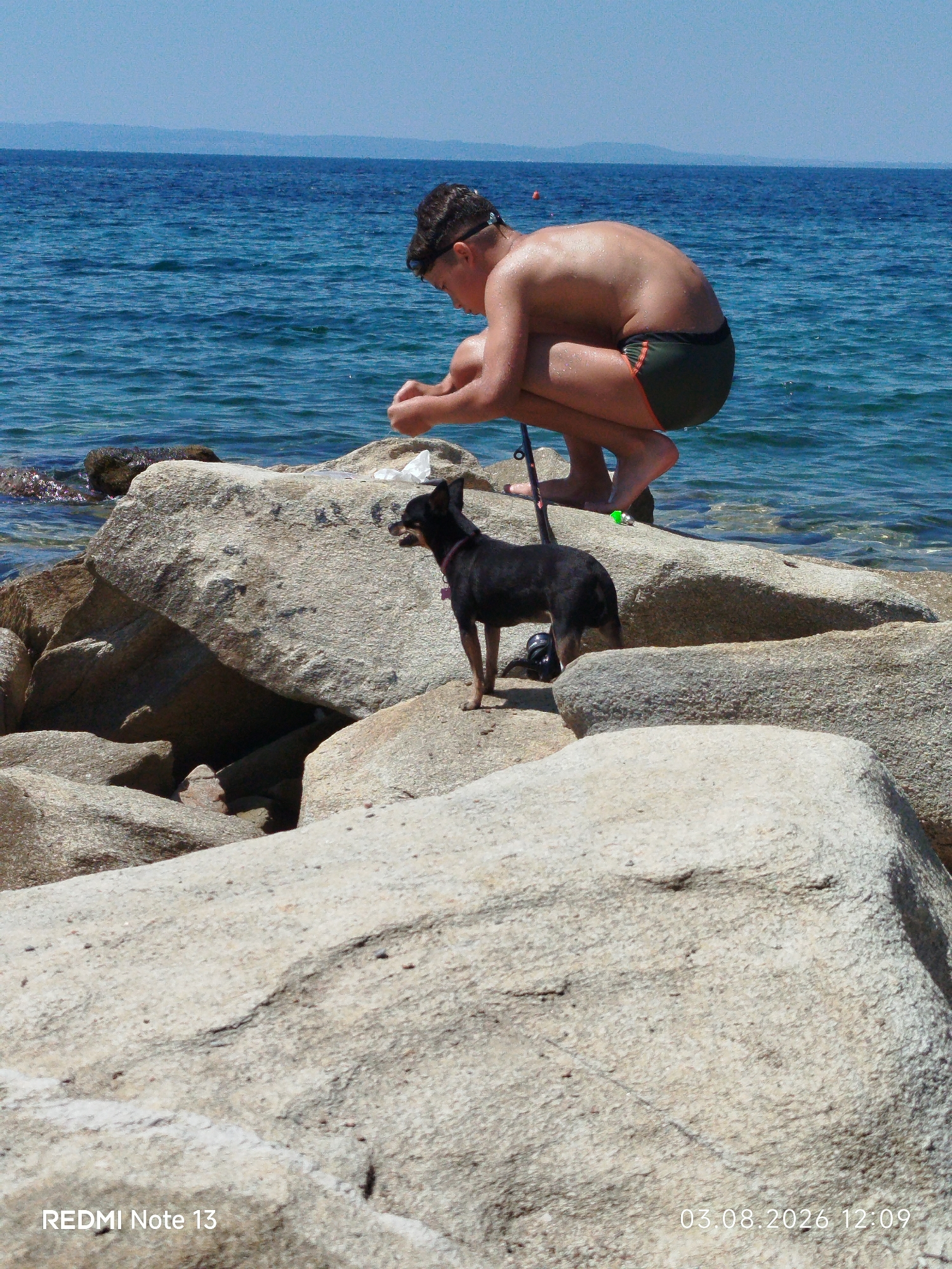 A young person is squatting on large rocks near the sea in Sithonia Municipal Unit, Greece, with a small black dog standing on the rocks beside them. The person is wearing dark swim trunks and has wet hair, suggesting they have been swimming or are near the water. They appear to be examining something in their hands. A fishing rod is placed vertically on the rocks near the person's feet. The dog is facing away from the person, looking towards the water. The environment is a rocky coastline with clear blue sea and a hazy coastline in the distance under a clear sky. The lower right corner of the image displays "03.08.2026 12:09" and the bottom left corner displays "REDMI Note 13".