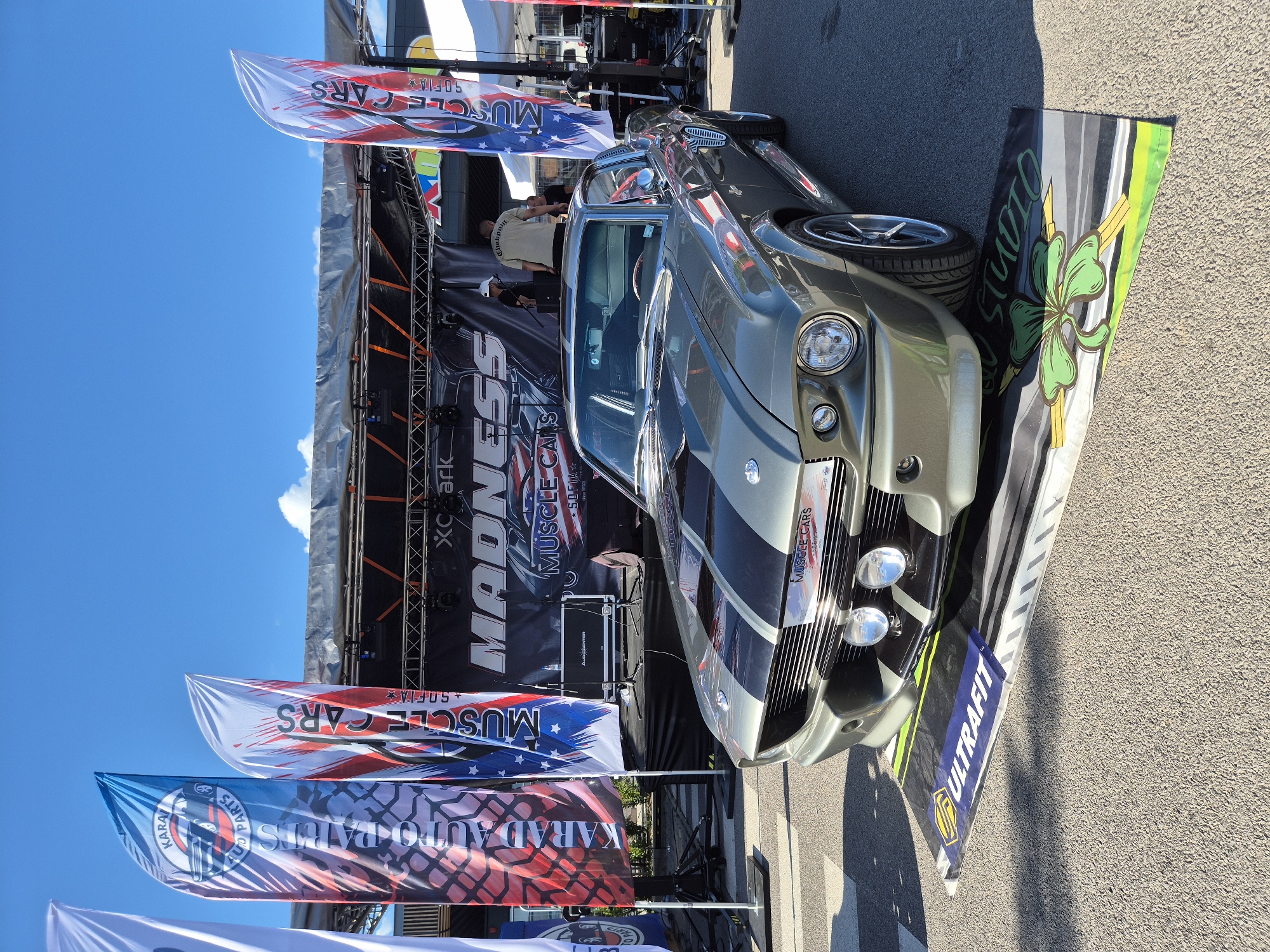 An outdoor, wide shot captures a grey 1960s Ford Mustang parked on an asphalt surface in front of a stage. The car is oriented facing right, with its front end prominent. The stage has a black backdrop with text that reads "MADNESS" and "MUSCLE CARS SOFIA". Various flags, some with "AUTO PARTS" and "MUSCLE CARS" text, are displayed on poles to the left of the stage. The sky is blue with some scattered white clouds. The scene appears to be an outdoor event or car show.
