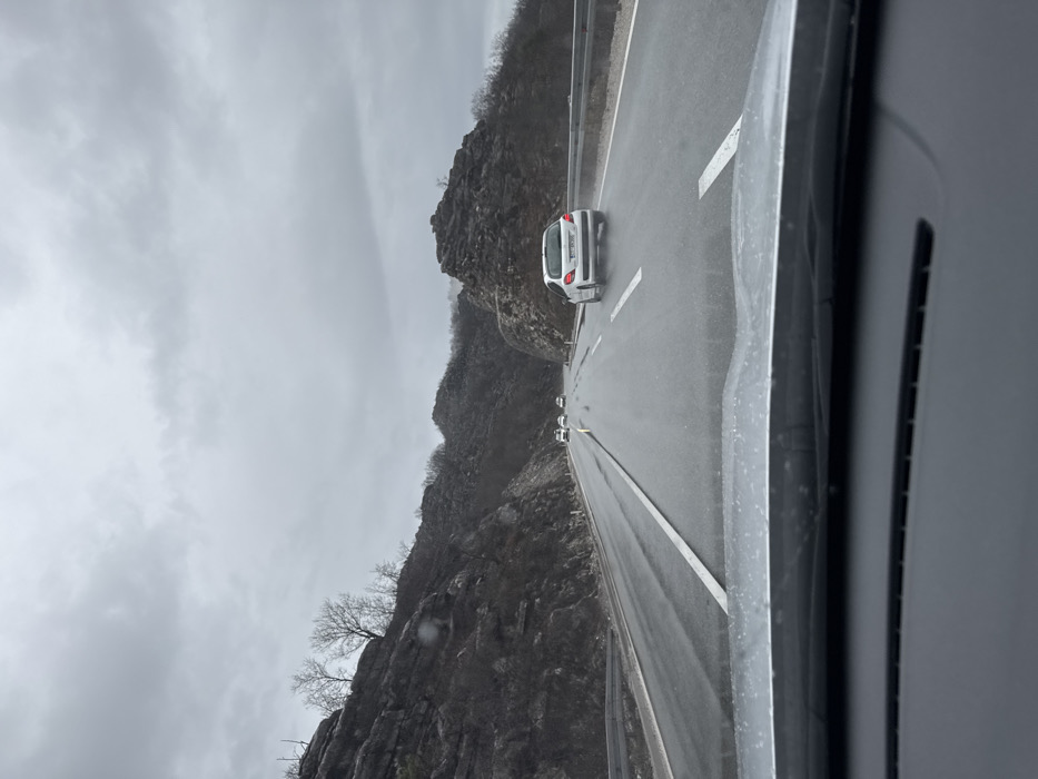 The image captures a wet, multi-lane highway in Gornji Ulići, Montenegro, viewed from the interior of a vehicle. The sky is uniformly overcast and grey. The road surface is visibly damp, with reflections. A white hatchback, identified by a partially visible Montenegrin license plate, is positioned in the right lane ahead of the camera vehicle, its rear tires generating a fine spray of water. Several other vehicles with illuminated headlights are present further down the road, receding into the distance. The highway is flanked on both sides by rocky, dark-colored hills, sparsely covered with leafless trees and dark brush. A metal guardrail runs along the right edge of the highway.
