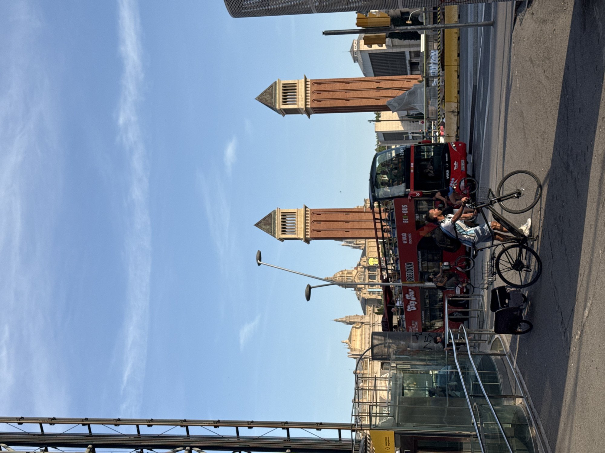 The image captures a street scene in Barcelona, Spain, featuring the iconic Venetian Towers of Montjuïc in the background. In the foreground, a man is riding a bicycle with a small trailer attached. A red double-decker "City Tour" bus is visible behind him, also carrying passengers. The scene appears to be a sunny day, with a clear blue sky overhead. The architecture suggests a European urban environment, with the distinctive towers serving as a key landmark. The overall impression is one of everyday urban life with a tourist element.