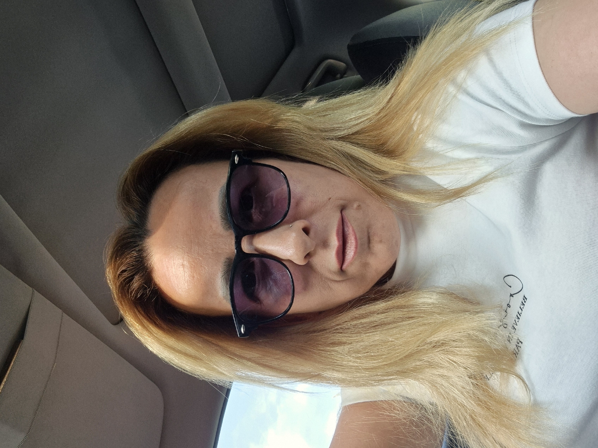 A woman with blonde hair and sunglasses is in a car. She is wearing a white t-shirt. The interior of the car is visible, including the ceiling and a seat. The background shows a glimpse of the sky and clouds through the car window.