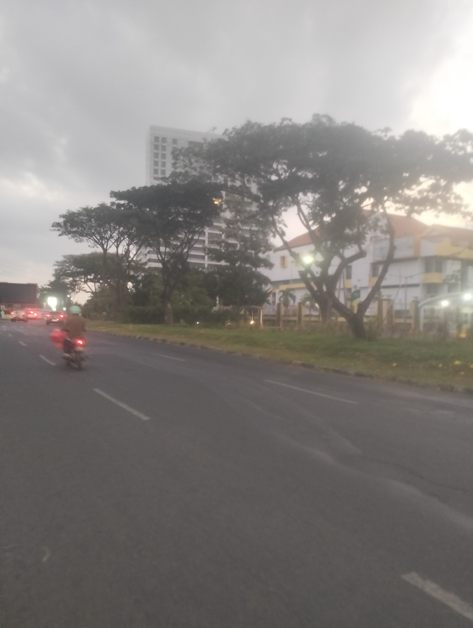 The image depicts a street scene in Surabaya, Indonesia, likely during overcast or twilight conditions. The perspective is from a moving vehicle, as indicated by the motion blur. In the foreground, a person on a motorcycle is visible, wearing a light brown jacket and a blue helmet, with red baggage attached to the back of the motorcycle. The motorcycle is in motion on an asphalt road marked with white lines. Further down the road, other vehicles with illuminated headlights are present.
In the background, several buildings are visible. A tall, modern white building stands prominently, with a grid-like pattern of windows. To the right of this, a lower, colorful building with orange and yellow accents is situated. Large trees with dense foliage line the road and partially obscure the buildings. The sky appears overcast, suggesting either an early morning, late afternoon, or a cloudy day. The overall impression is a typical urban street environment in Surabaya.