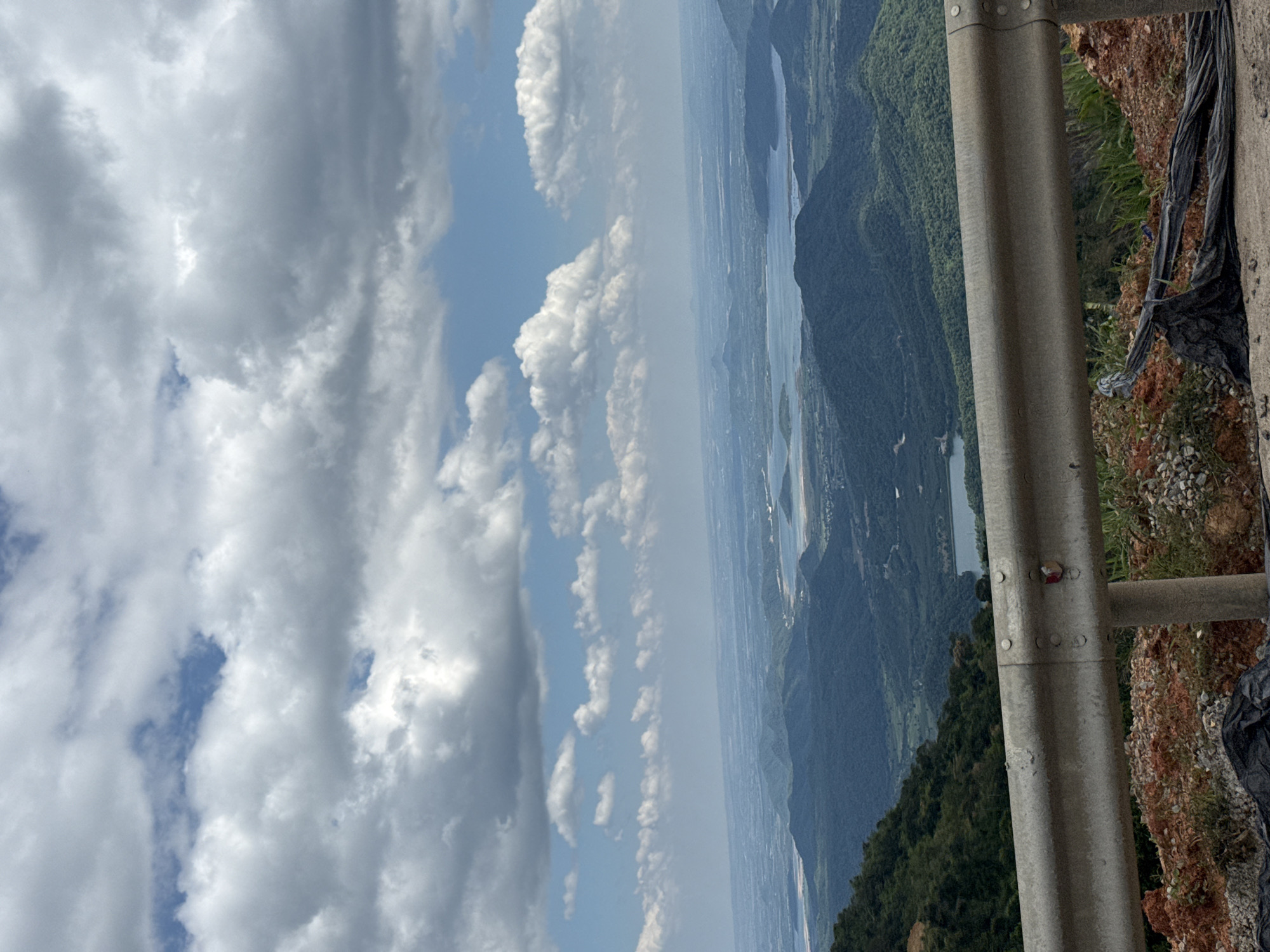 The image depicts a scenic overlook in Nam Hai, Vietnam, with a vast expanse of rolling green hills and a large body of water in the distance. The foreground features a metal guardrail, indicating a roadside viewpoint, with loose gravel and dirt on the ground. The sky above is filled with a dramatic display of white, fluffy clouds against a bright blue backdrop. The overall impression is one of natural beauty and a peaceful, expansive landscape.