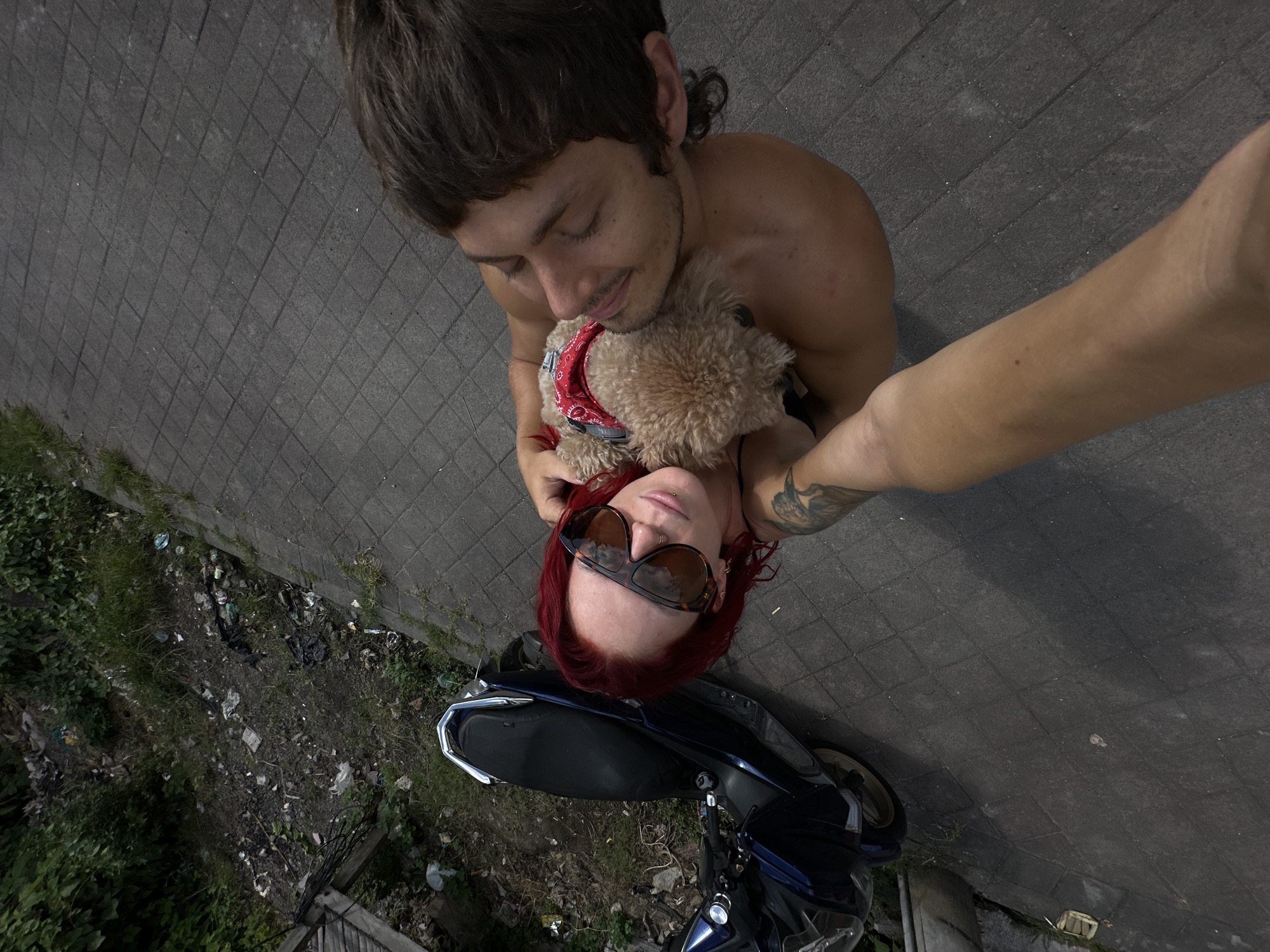 The image is a selfie taken from an elevated angle, featuring a man, a woman, and a small dog. The man, shirtless, has brown hair styled in a mullet. He is hugging a small, tan-colored dog wearing a red bandana. The woman, with red hair and wearing large sunglasses, is positioned below him.
They are standing next to a blue scooter parked on a grey brick surface. Adjacent to the brick surface, there is a strip of overgrown greenery with scattered trash.
Based on visual cues of the location, it is the city of North Kuta, Indonesia.