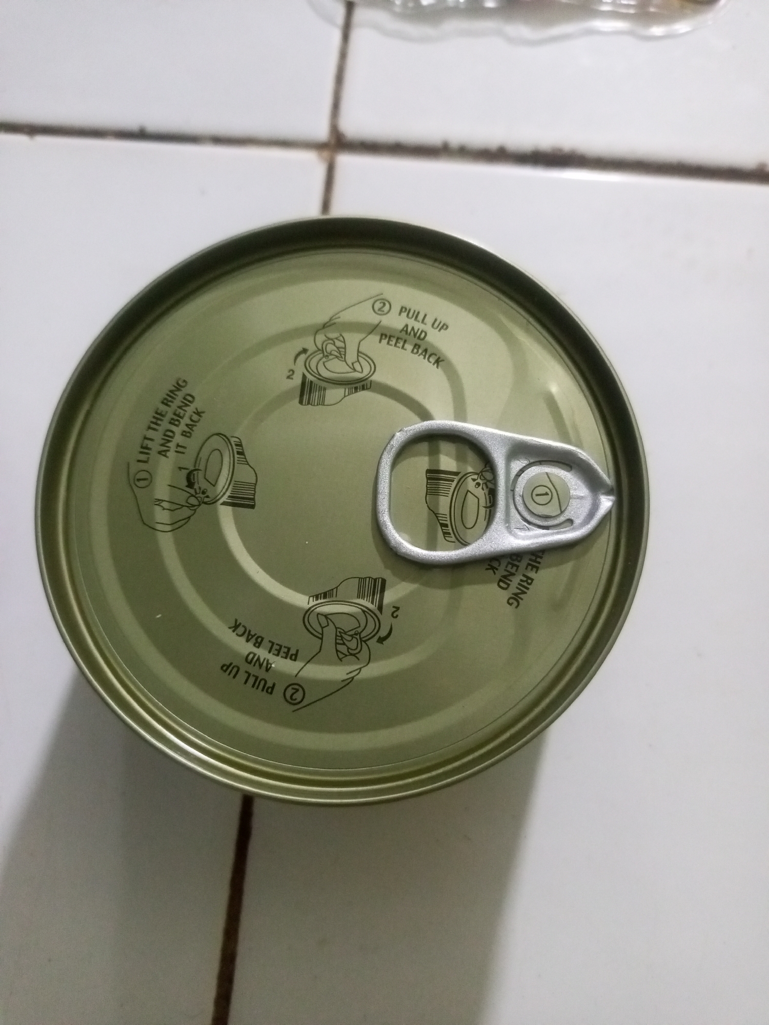 A top-down view shows the lid of a green metal can on a tiled surface. The can lid features a pull tab and etched instructions for opening. These instructions are illustrated and text-based, showing two steps for opening. One set of instructions is oriented to be read from the top left, with text including "LIFT THE RING AND BEND IT BACK." Another set of instructions is oriented to be read from the bottom right, with text including "PULL UP AND PEEL BACK." The tiled surface appears to be a floor with white tiles and dark grout lines. The lighting suggests an indoor setting, possibly a kitchen or pantry. There are no people visible in the image. The scene is static. The location is not specified as being in La Tagua, Venezuela.