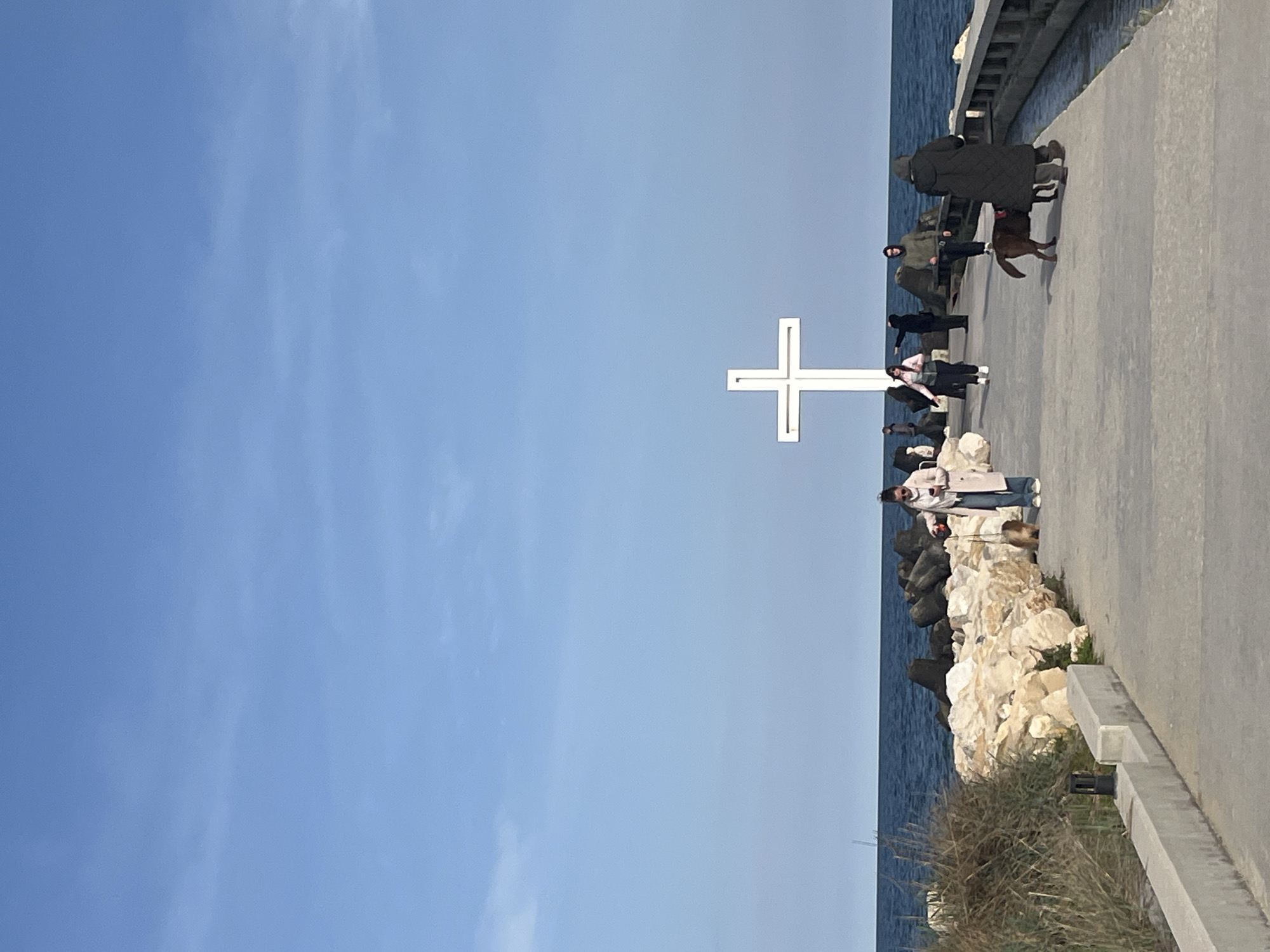 The image depicts a scene in Varna, Bulgaria, featuring a large white cross situated on a breakwater along the Black Sea. A group of people stroll along a paved pathway running alongside the breakwater. Some are observing the sea while others appear to be walking their dog. The sky is clear and blue. The breakwater is constructed from large white rocks. The image is taken on a sunny day.
