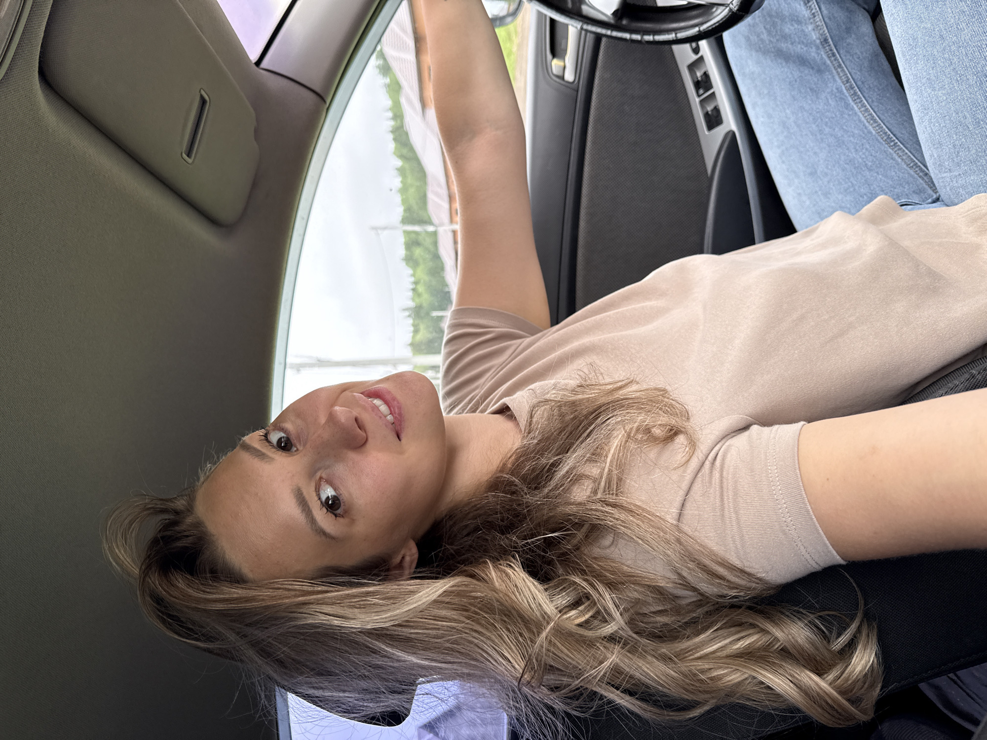 A woman with long, wavy blonde hair is sitting in the driver's seat of a car, looking at the camera and smiling. She is wearing a light brown t-shirt and blue jeans. The interior of the car is visible, including the steering wheel, dashboard, and a sun visor. The outside of the car shows a blurred background of houses and trees under an overcast sky, suggesting an outdoor setting in a rural or suburban area. The image appears to be taken on a cloudy day. The location is identified as the city of Chaikovsky, Russia.