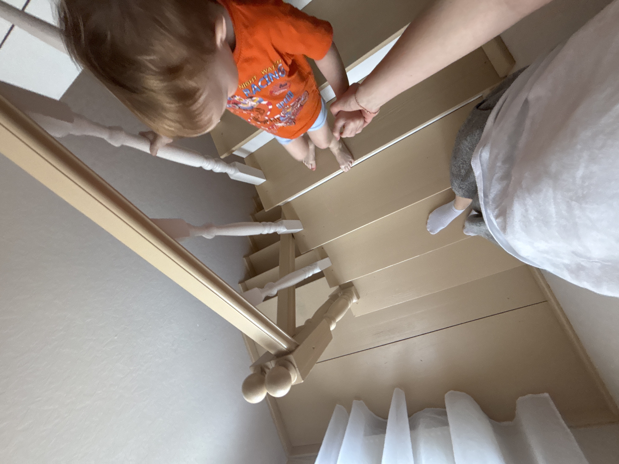 A person assists a child down a flight of painted wooden stairs. The child wears an orange t-shirt and blue shorts, holding the hand of the adult and the railing. The adult wears grey pants, a white shirt, and a white sock on their visible foot. The stairs have white balusters. White curtains are visible at the bottom of the stairs. The scene is indoors. The location is Ферма, Russia.