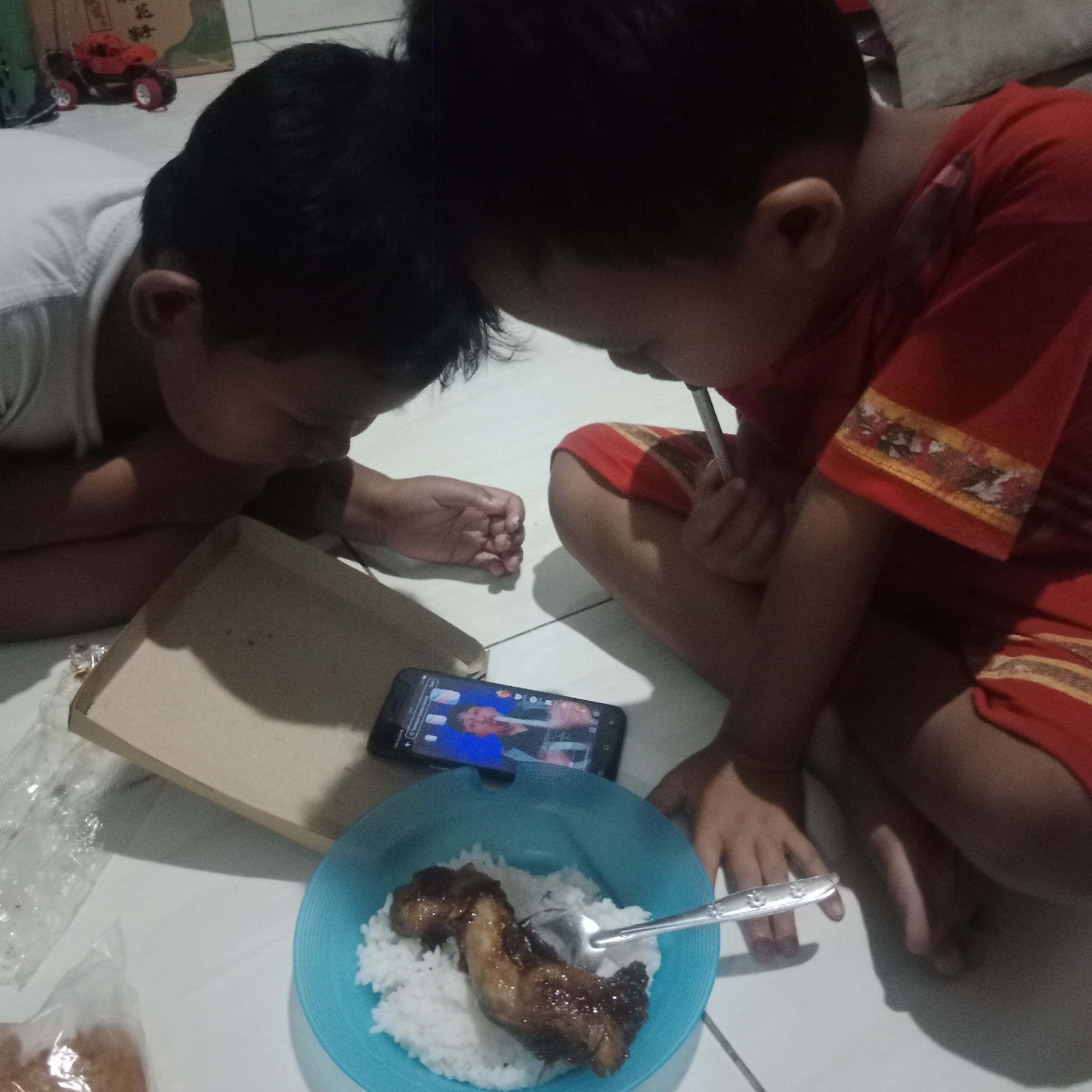 Two young boys are positioned on the floor in what appears to be a domestic setting, possibly in Tegowanu Kulon, Indonesia. The boy on the left, wearing a white shirt, is leaning forward, with his head bowed towards a brown cardboard box. His right arm is extended, resting on the box. The boy on the right, wearing a red shirt with decorative trim, is also leaning forward, looking at a smartphone placed on the floor. He is holding a utensil, possibly a straw, in his right hand. In the foreground, a blue bowl contains white rice topped with a piece of meat. A spoon rests in the bowl. To the left of the bowl, a clear plastic bag contains food items. In the background, a red toy truck is visible, along with packaging with text in what appears to be an Asian language. The overall lighting suggests an indoor environment.