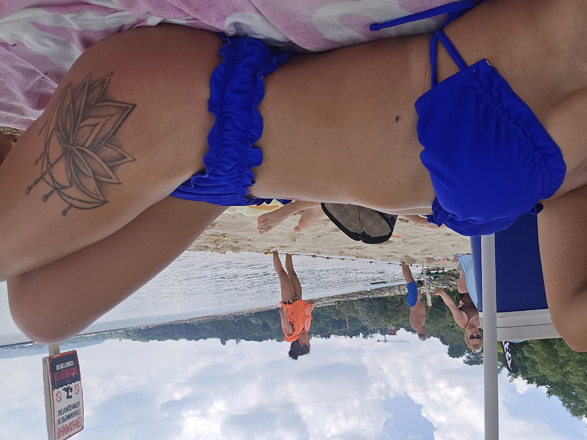 The image depicts a beach scene at Varna, Bulgaria, likely during daytime. In the foreground, a person is lying on a pink and white striped towel on the sand, wearing a blue bikini. A portion of their legs and torso is visible, with a tattoo of a flower on their thigh. Behind them, other beachgoers are visible. A woman with blonde hair pulled up is standing near a cooler, wearing sunglasses and a brown swimsuit, interacting with a man in blue shorts. Further in the background, a child in an orange life vest stands near the water. The sea is calm, and there is a tree-lined coast in the distance, with some construction cranes visible on the left. A warning sign with text in a foreign language is posted on the right. The sky is partly cloudy.
