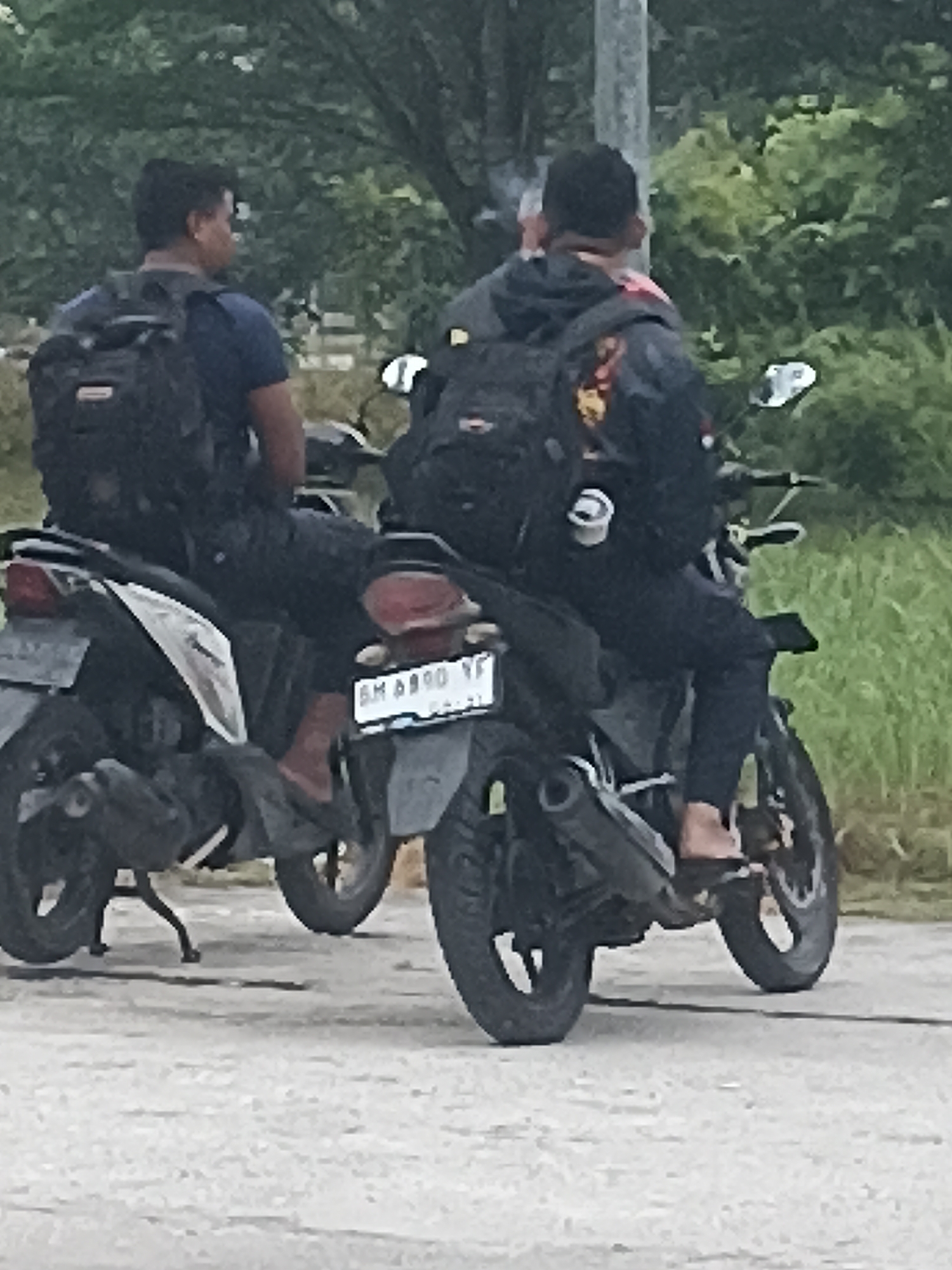 Two individuals are seated on motorcycles, facing away from the camera, on a paved surface in Kampung Tengah, Indonesia. The individual on the left is wearing a dark blue t-shirt and black pants, with a black backpack. The motorcycle they are on is silver and black. The individual on the right is wearing a dark jacket with a design on the back and dark pants, also with a black backpack. Their motorcycle is dark colored and has a visible license plate. The background consists of green foliage and trees, suggesting an outdoor environment. The lighting appears to be overcast.