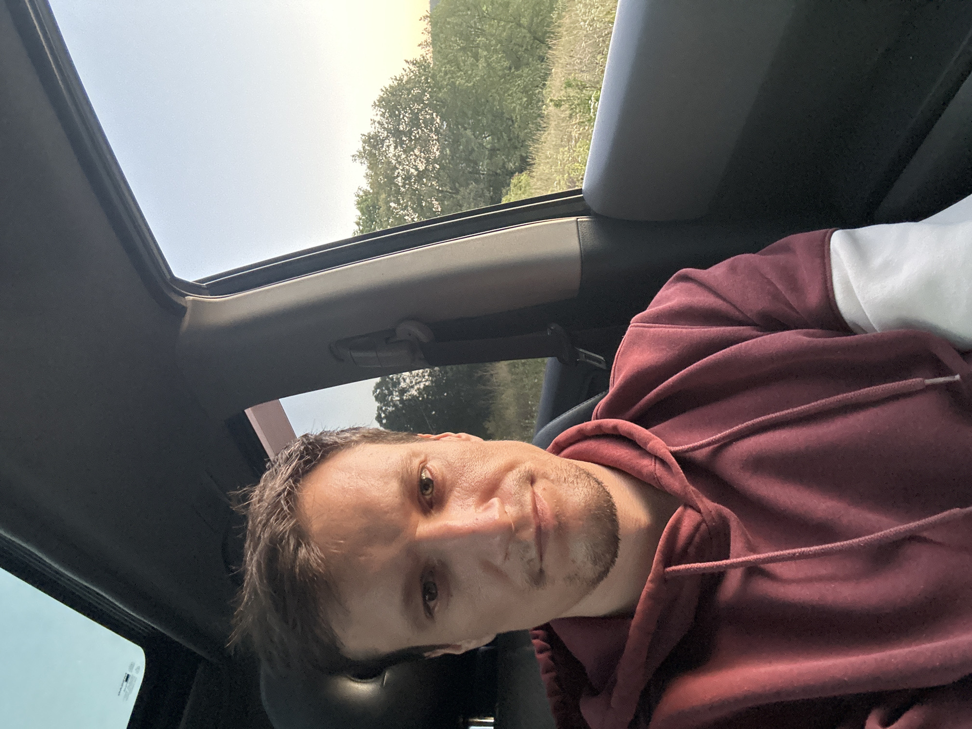 A man with light brown hair and stubble is sitting in the passenger seat of a car, looking directly at the camera. He is wearing a maroon hoodie with white accents on the sleeves. The car's interior is visible, including the headliner, a sun/moon roof, and part of the dashboard and door. Through the open passenger window, a rural landscape with trees and tall grass is visible under a clear, pale sky, suggesting it is either dawn or dusk. The scene is consistent with a location on the outskirts of Makiv, Ukraine, a rural village.