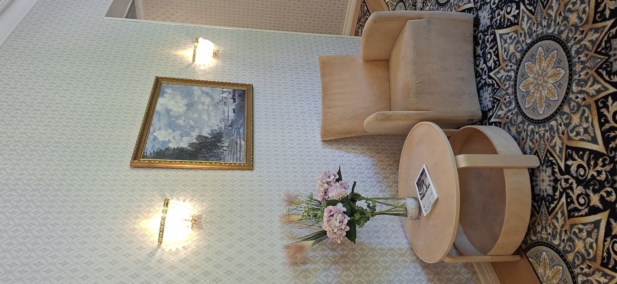 A still life photograph depicts an interior scene with furniture, art, and floral arrangements, set against a patterned wallpaper. The image captures a corner of a room, featuring a light beige armchair on the right, upholstered in a soft fabric. Adjacent to the armchair, to the left, is a round, light-colored wooden coffee table with a lower shelf. On the table, a glass vase holds a bouquet of pale pink hydrangeas and some dried ornamental grass. A magazine with a visible title of "GUIDE" lies open on the table, partially obscured by the floral arrangement. Above the table and armchair, a framed painting hangs on the wall. The painting is a landscape depicting a body of water with boats, bordered by trees on one side, under a cloudy sky, and framed in gold. Two wall sconces with crystal accents illuminate the space, one to the left of the painting and another to its right. The wallpaper has a subtle, repeating geometric pattern in a light, neutral tone. The floor is covered by a dark rug with an intricate, ornate pattern in gold, black, and grey. The scene does not include any people or animals. The style suggests a well-maintained living space or waiting area. The specific location in Lahti, Finland is not discernible from the visual information provided in the image.