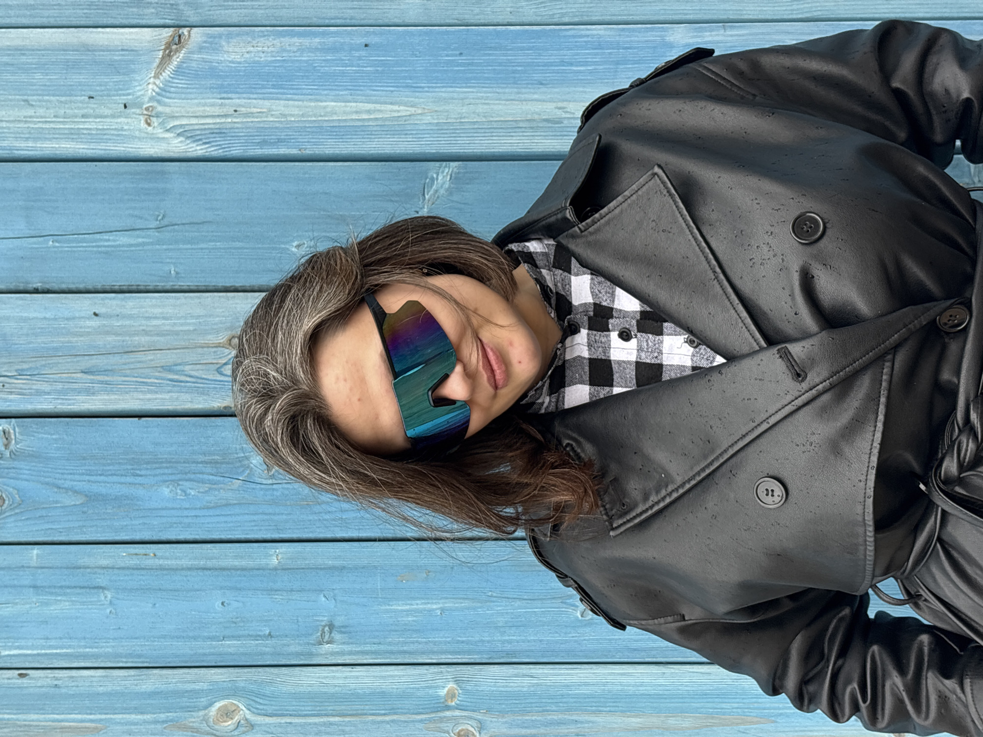 A woman with shoulder-length gray and brown hair is wearing large, futuristic-looking sunglasses. She is also wearing a black-and-white checkered shirt under a black leather trench coat, which is buttoned. The background consists of light blue horizontal wooden planks. The image appears to be taken in Chaikovsky, Russia.