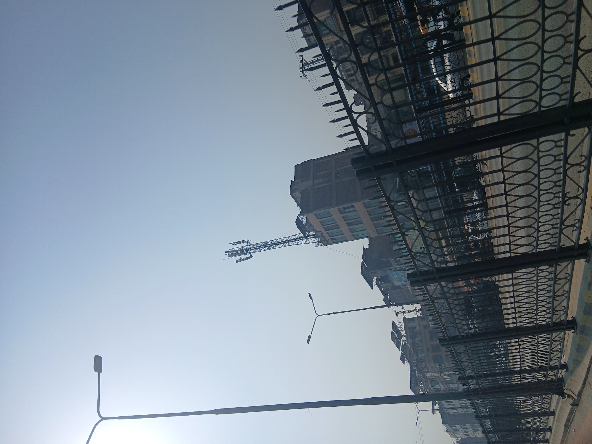 The image depicts a street scene in Peshawar City Tehsil, Pakistan, under a bright, hazy sky. A tall, dark lamppost stands on the left side, partially obscuring a faint hint of sunlight. In the background, several buildings of varying heights are visible, some appearing older with weathered facades, while others are more modern. A prominent telecommunications tower with multiple antennas rises above one of the buildings. A decorative black metal fence with ornate scrollwork lines the front of the scene, separating the street from what appears to be a sidewalk or pedestrian area. Beyond the fence, a road is visible with a few vehicles. The overall impression is of an urban environment on a clear, possibly warm, day.