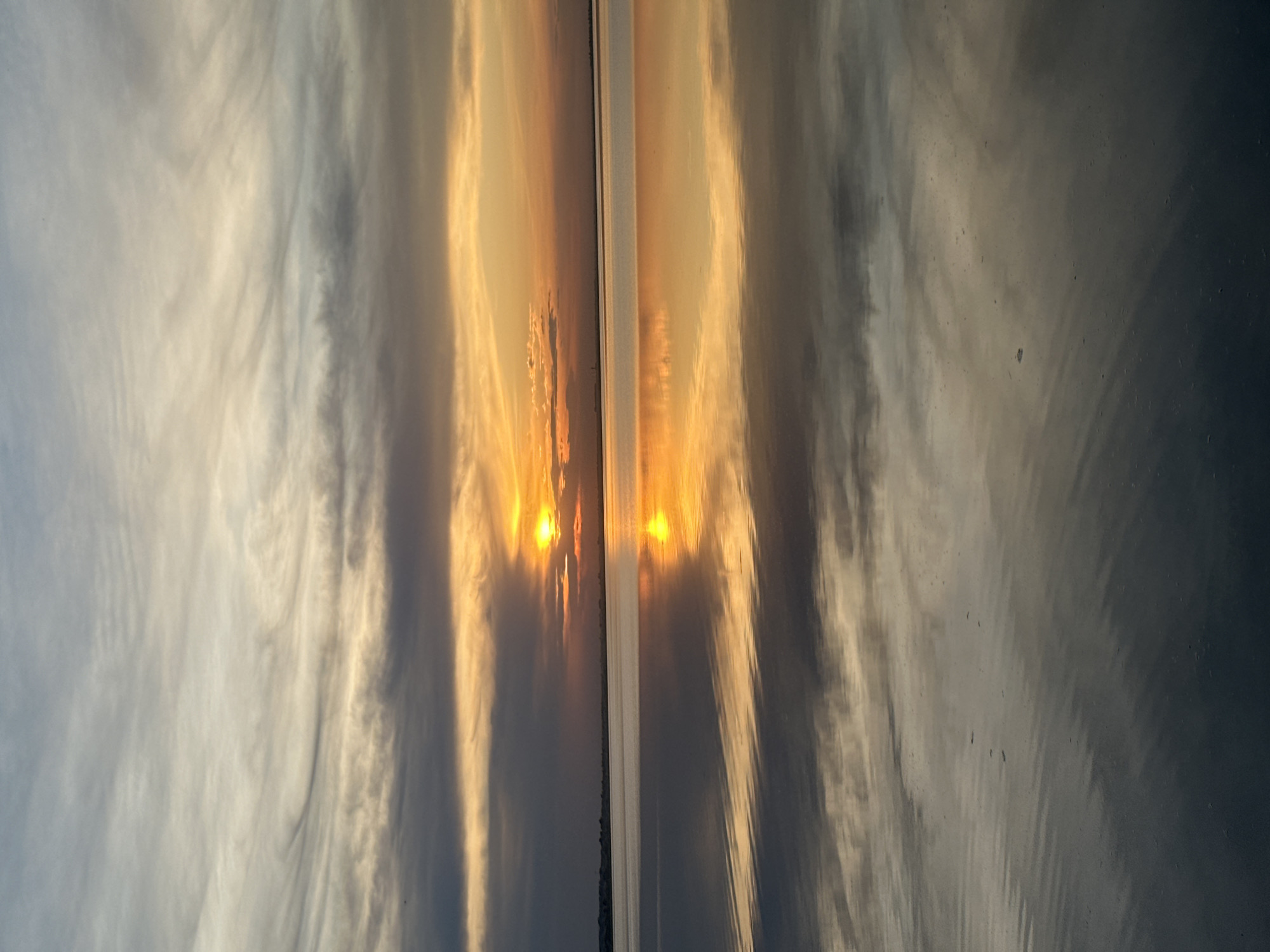 The image shows the reflection of a sunset on a wet surface. The sun is partially visible, reflecting a bright orange glow. Clouds are visible in the background and their reflection creates a swirling pattern on the wet surface. The location appears to be near a body of water, possibly a lake, in Kokshetau, Kazakhstan.
