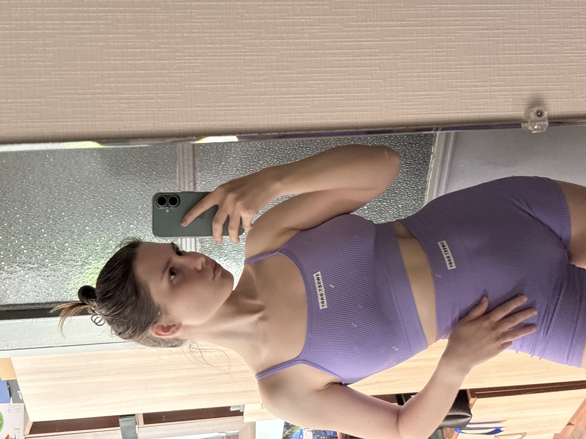 A woman is posing for a selfie in front of a mirror. She is wearing a light purple, ribbed workout set consisting of a sports bra and shorts. Her brown hair is pulled back into a messy bun. The mirror reflects a room with a light-colored, horizontally-patterned wall and a window with textured glass. The objects and setting details are insufficient to geolocate the image to Saint Petersburg, Russia.
