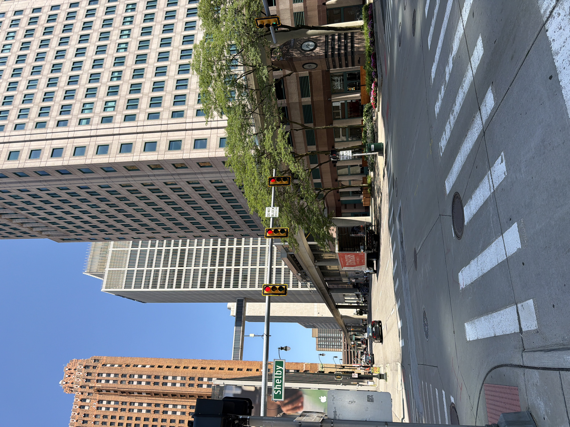 The image depicts a street scene in the city of Detroit, United States of America, likely in the downtown area. Towering skyscrapers dominate the skyline. On the left, a distinctive copper-colored Art Deco building with numerous windows stands prominently. To its right, a modern, light-colored skyscraper with a repeating grid of windows rises. Further to the right, another large, light-colored skyscraper with a similar window pattern is visible.
In the foreground, a street intersection with crosswalks is apparent. A traffic light is visible, showing a red signal. A street sign reads "Shelby." A train track system, suspended above the street by concrete pillars, runs horizontally across the mid-ground. A red delivery truck with "Red" and "Soaring" visible on its side is parked on the right side of the road. Several cars are driving on the street. Trees with green foliage are planted along the sidewalk. The sky is clear and blue.
