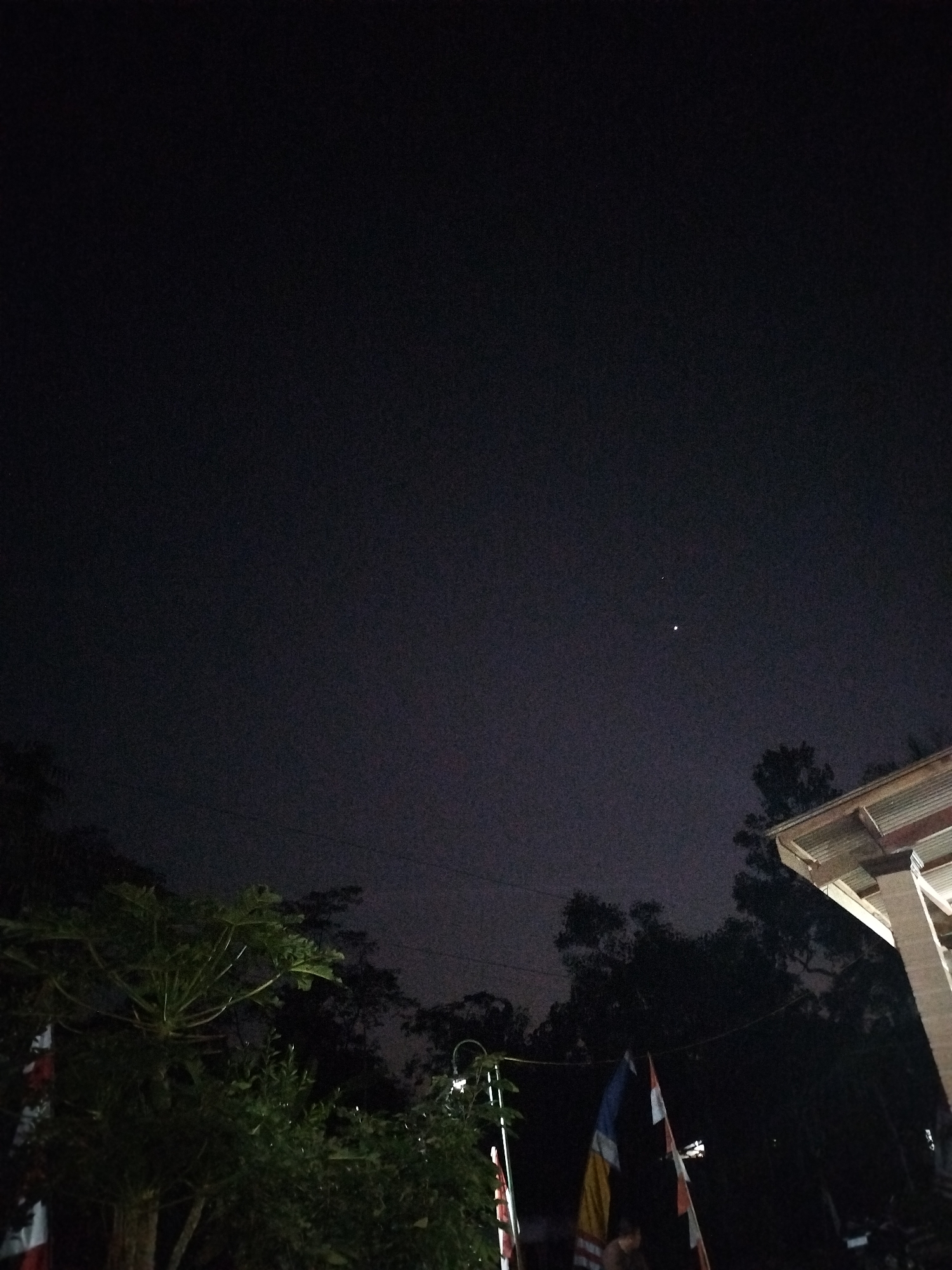 The image depicts a nighttime scene in Sewurejo, Indonesia, with a dark, cloudy sky. Two celestial bodies are visible in the sky, likely stars or planets. In the foreground, there are dark trees, a building with a corrugated roof, and several flags. One flag appears to be red and white, possibly the Indonesian flag, while others are blue, yellow, and white. The lighting is dim, suggesting it is nighttime. There are no people clearly visible in the image. The environment appears to be an outdoor area, possibly near a residential or public space, given the presence of flags and a building. The overall impression is one of a quiet, nocturnal setting.