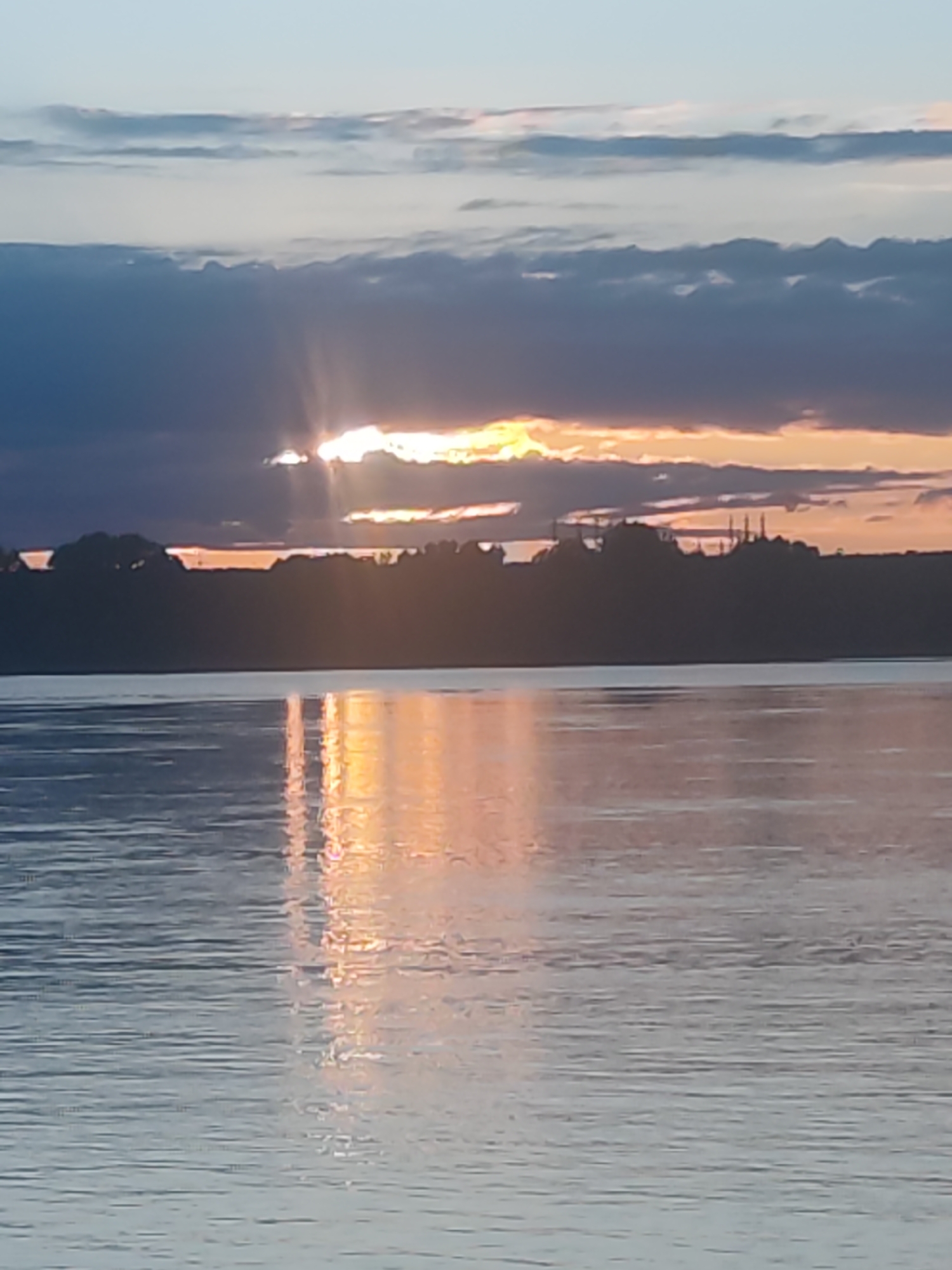 The photograph captures a stunning sunrise or sunset over a vast body of water, likely a river or lake, within the geographical context of Noviy, Russia. The scene is dominated by a dramatic sky and its reflection on the water.
The sun, positioned low on the horizon, is partially obscured by a band of dark, heavy grey and blue clouds that stretch horizontally across the mid-sky. Radiant beams of golden and orange light pierce through and emanate from behind these clouds, creating a powerful visual effect with distinct sunrays. The edges of these dark clouds are illuminated with warm orange and yellow hues where they catch the sun's light. Above this darker cloud layer, the sky transitions to a lighter, ethereal blue-grey with softer, more diffuse clouds.
The intense golden and orange light of the sun is vividly reflected on the surface of the water, forming a broad, shimmering path that ripples towards the viewer. This reflection creates a dynamic contrast with the cooler blue and grey tones of the water away from the sun's direct glare. The water's surface shows gentle ripples and textures, indicating a calm environment.
On the distant horizon, a dark, silhouetted landmass, possibly a forested area or low hills, stretches across the frame. Faintly visible against the brighter sky on the right side of this silhouette are several slender, pointed structures, resembling minarets or distant spires, adding a subtle architectural element to the natural vista. There are no people, animals, or discernible text visible in the image. The overall impression is one of tranquil grandeur and natural beauty, emphasizing the dramatic interplay of light, clouds, and water at dawn or dusk.