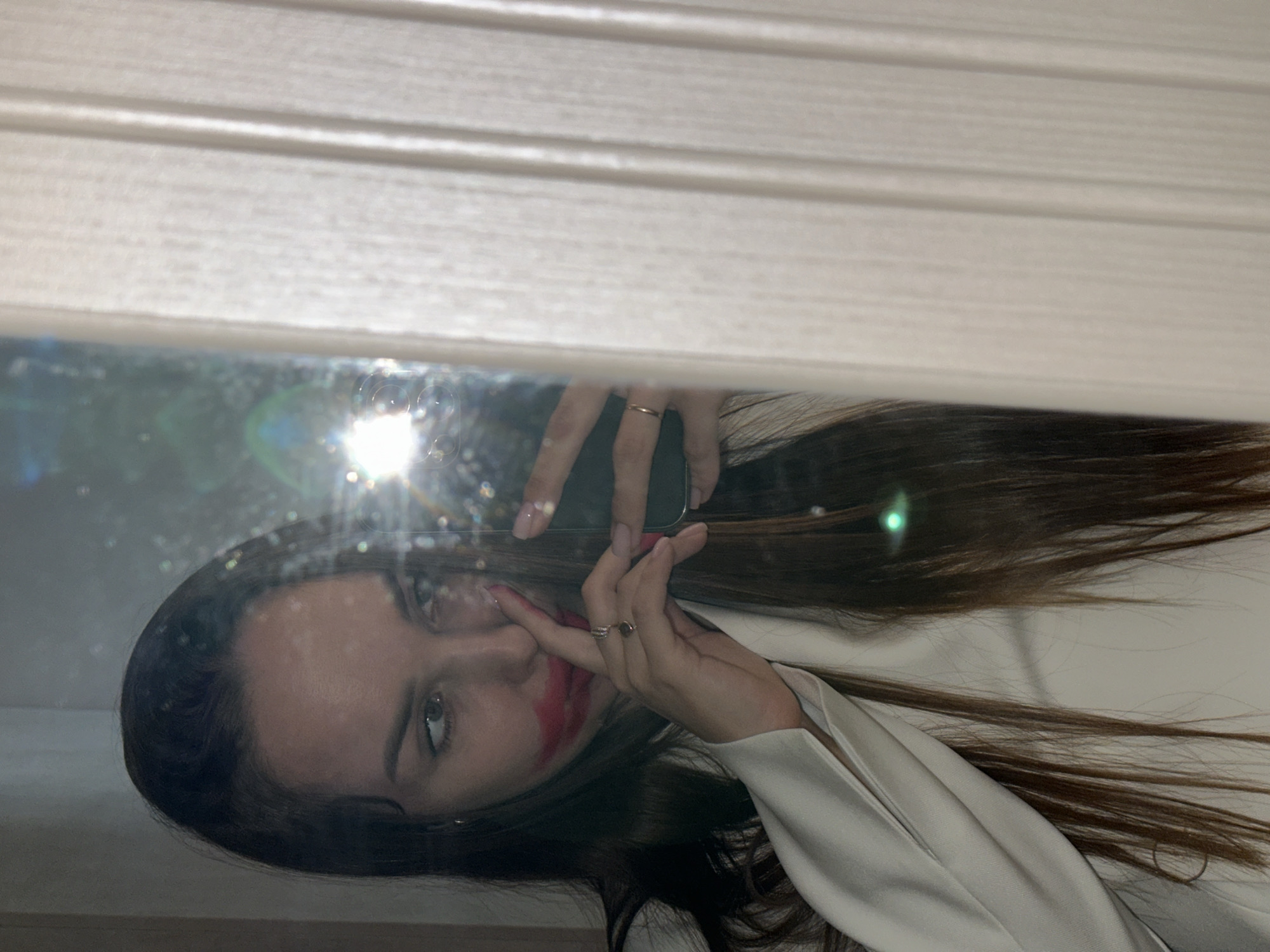 A young woman is reflected in a mirror. She is holding a smartphone above her head, presumably to take a selfie. The woman has long brown hair and is wearing a white garment, possibly a jacket or blouse. Red makeup or lipstick is smeared around her mouth and cheek. The mirror is partially obscured by white horizontal blinds above it. The scene's location and whether it's in the city of Chaikovsky, Russia, cannot be determined from the image.