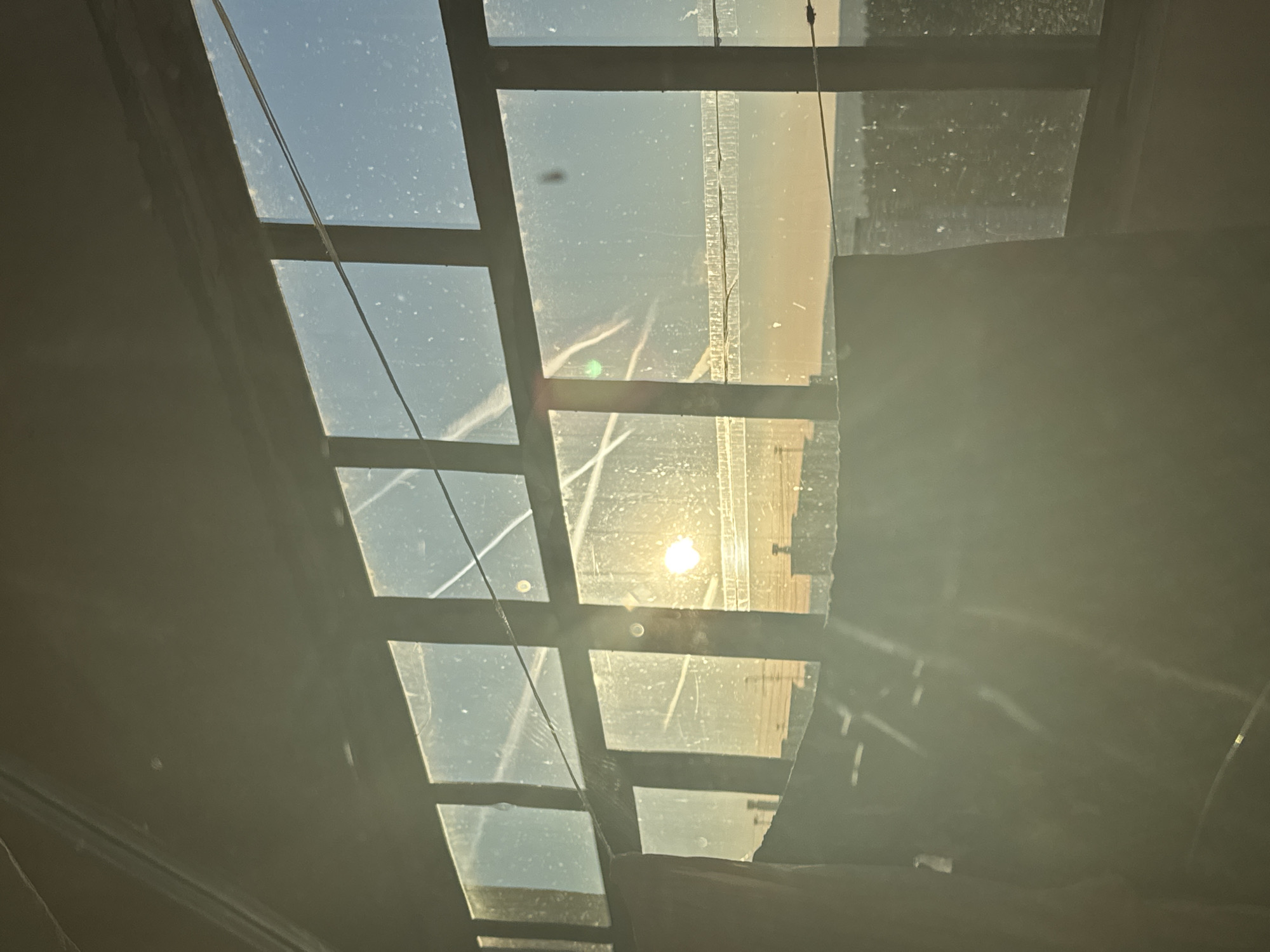 The image depicts a view looking up through a multi-paneled window or skylight. The sun is visible through one of the panes, casting a bright light and shadows. The panes of glass are dirty, with some streaking and residue visible. There is a grid-like structure made of wooden frames, most likely supporting the glass panes. The scene appears to be inside a building, given the framing structure and enclosed nature. The surrounding environment is dimly lit. The image does not provide information to confirm it was taken in Chaikovsky, Russia.