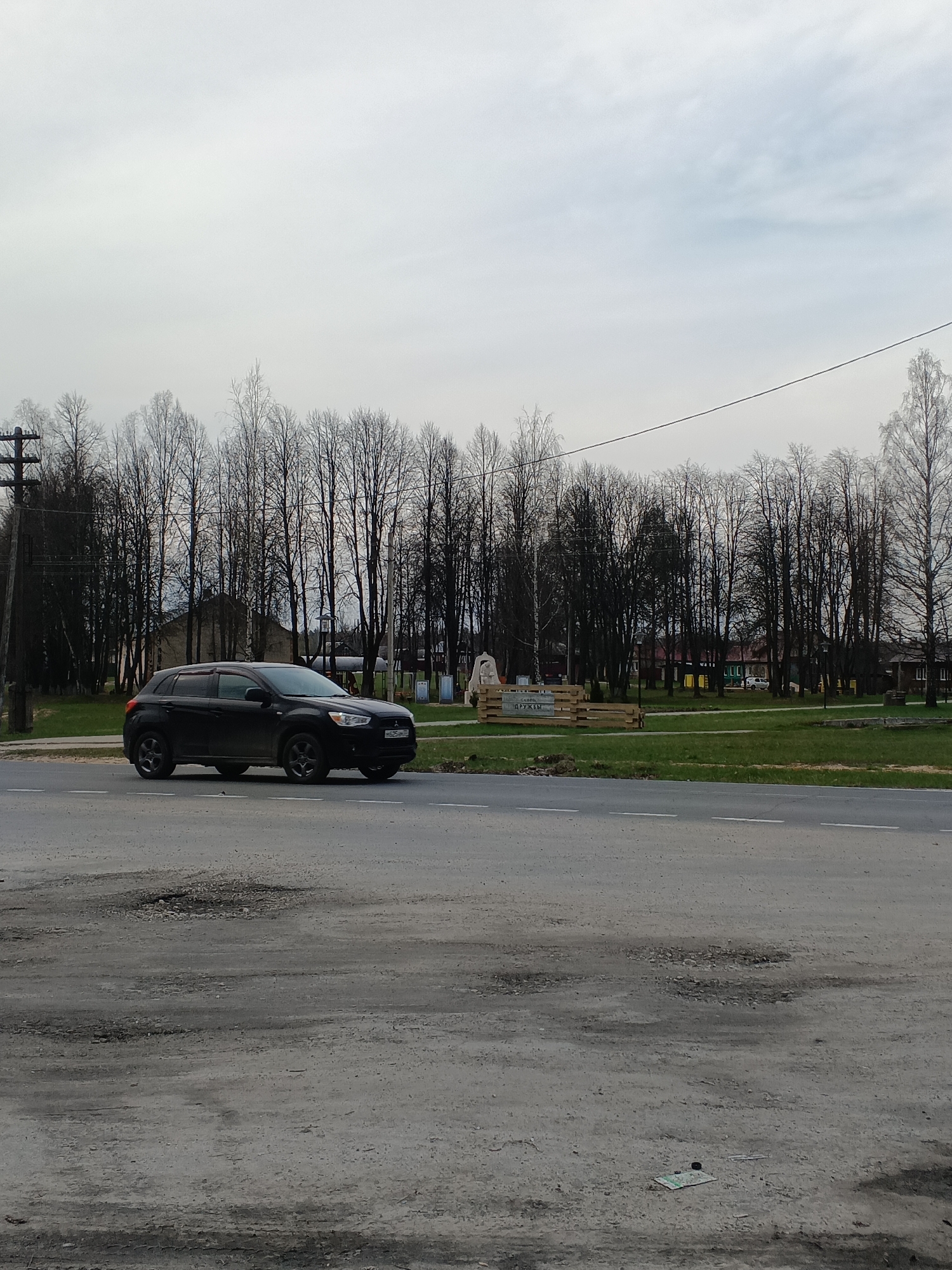 A black SUV is parked on a paved road with a white lane divider in the city of Красный Маяк, Russia. The foreground is a concrete surface with potholes and debris. Beyond the road is a grassy area with trees and some buildings in the distance. A utility pole is visible on the left side of the image. A wooden structure is located on the grass behind the SUV. The sky is overcast.