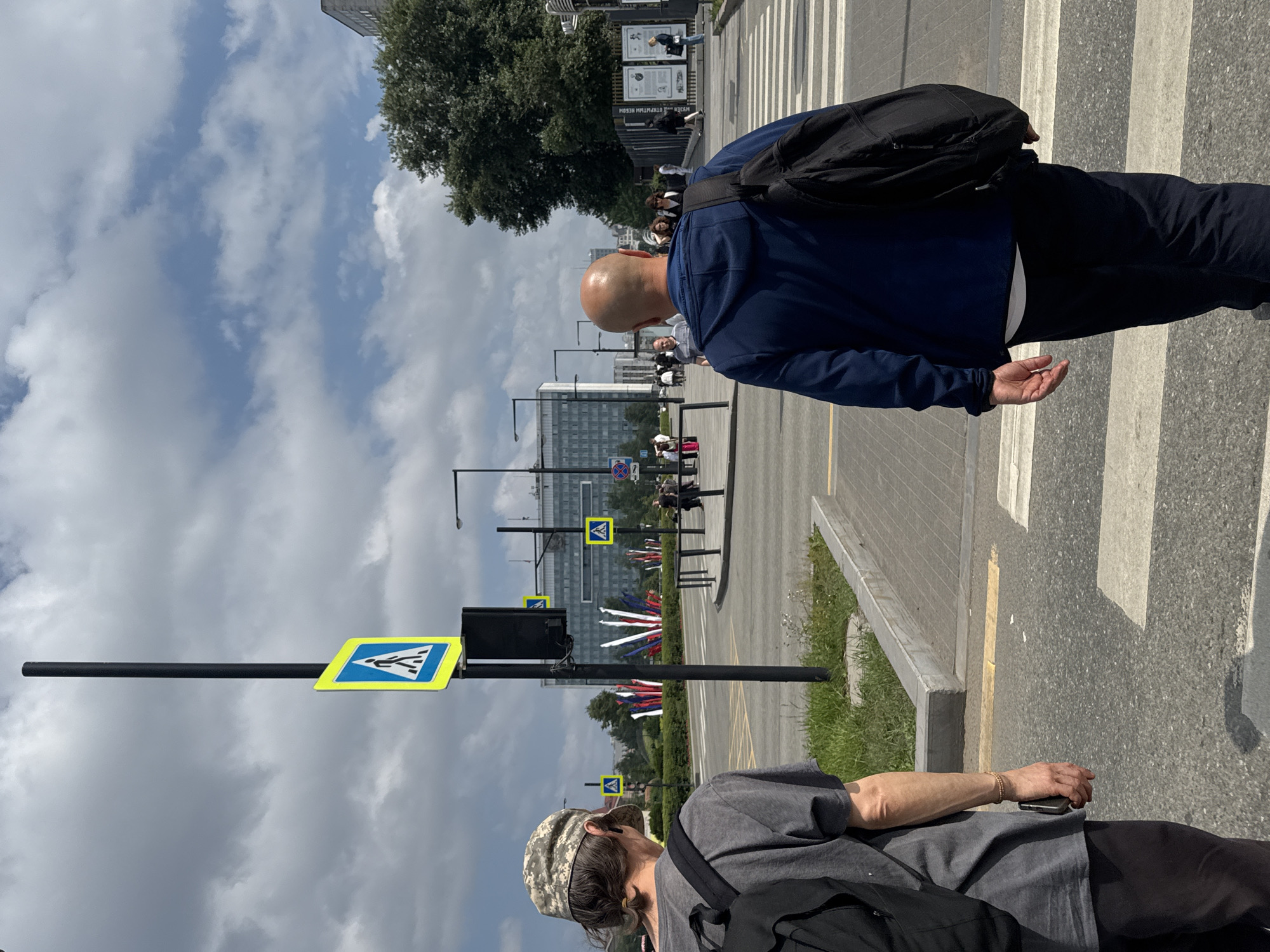 Two men are walking across a pedestrian crossing in Perm, Russia. The man on the left, facing away from the camera, is wearing a grey t-shirt, black pants, and a camouflage baseball cap. He has a black backpack on. The man on the right, also facing away, is wearing a blue hoodie, dark pants, and has a black backpack. In the background, there are several buildings, trees, and pedestrian crossing signs. The sky is partly cloudy.