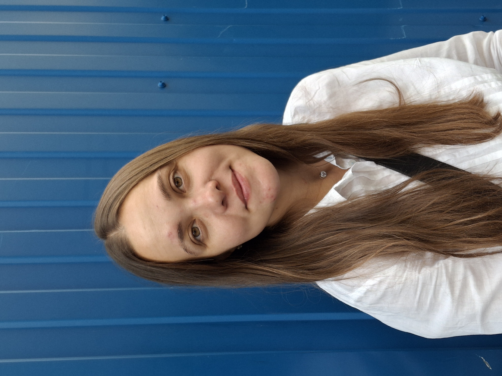A medium shot portrait of a young woman is captured against a vibrant blue corrugated metal wall. The woman has long, straight brown hair that falls past her shoulders and is parted in the middle. She is wearing a white collared shirt, and a black strap is visible across her chest. She has a small, silver-colored pendant around her neck. Her facial features are visible, and she has a gentle smile, with her head tilted slightly to her left. The background wall has vertical ridges and a few visible circular fasteners. The lighting suggests an outdoor setting. The scene is likely in the city of Chaikovsky, Russia, given the specific architectural style of the wall.