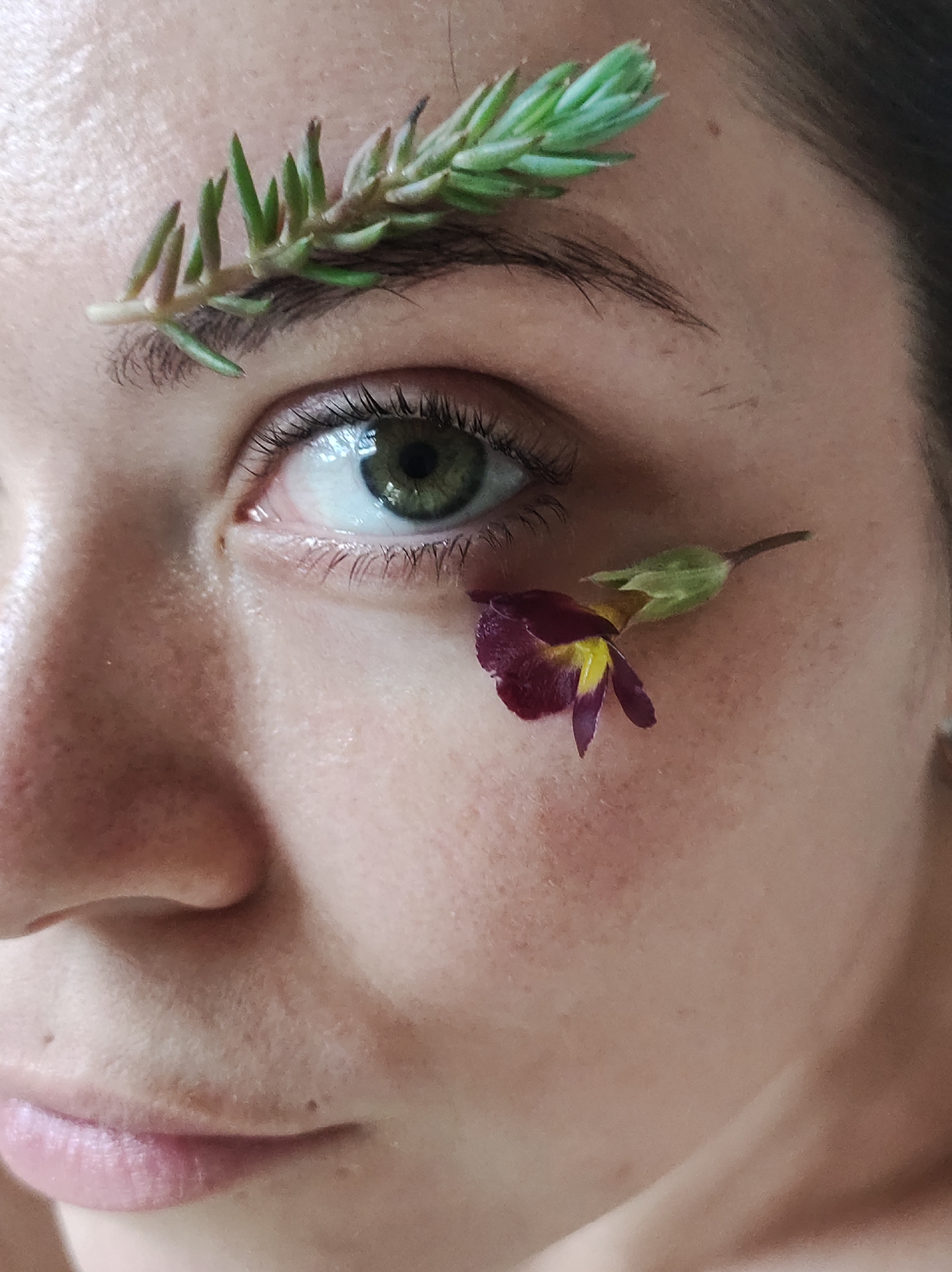 A close-up shot focuses on a person's face, specifically around their left eye. A sprig of green succulent-like leaves rests on the eyebrow. Below the eye, a small, deep red flower with a yellow center and attached green bud is positioned on the cheek. The person's eye is green, and their skin exhibits a natural texture with visible pores. The image is well-lit, highlighting the details of the face and the botanical elements. No location information is contained in the visual content.
