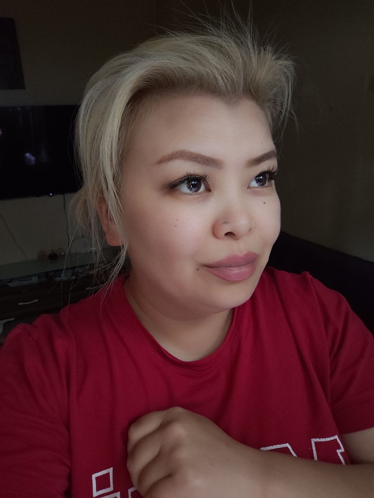 A medium indoor shot depicts a woman with fair skin and platinum blonde hair, wearing a red t-shirt with white blocky patterns on the lower right. She has her right hand up, near her chest. The woman has light eyebrows, dark eyelashes, and a small nose piercing. Her lips are adorned with a pinkish nude lipstick. She is looking slightly upwards and to her right. Behind her, a dark-colored TV is faintly visible, suggesting the location is likely a living room or bedroom in the city of Unknown, Unknown. The background is dark, with a blurry indistinct object behind the TV.