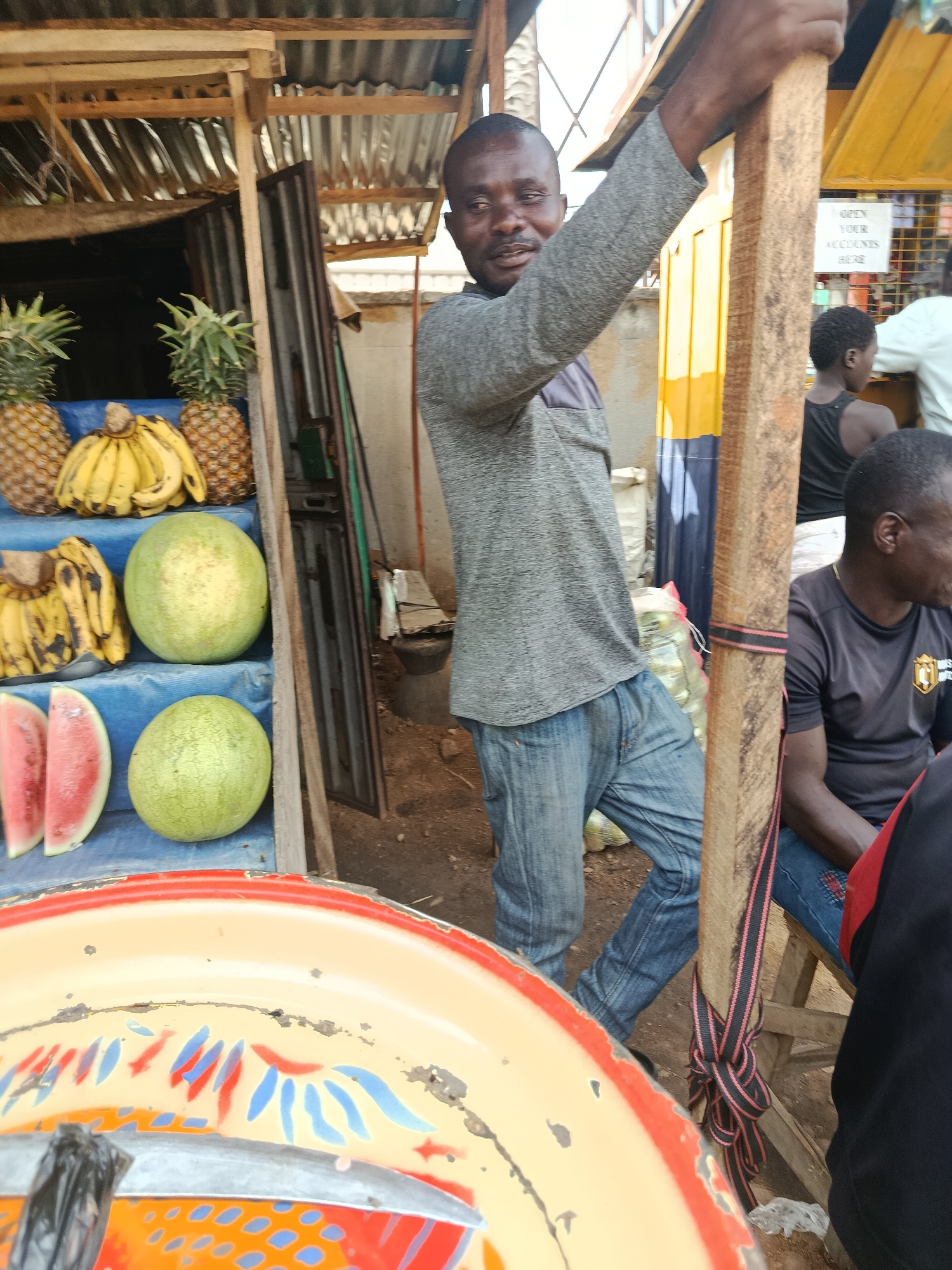 The image shows an outdoor market scene in Jos, Nigeria, focusing on a fruit stall and the people present. A man, appearing to be a vendor, stands to the right of the frame, holding onto a wooden post. He is wearing a gray long-sleeved shirt and blue jeans. Behind him, a selection of fruits is displayed, including pineapples, bananas, watermelons, and what appear to be two slices of watermelon in the foreground. To the right of the main vendor, another person is seated, wearing a dark t-shirt with a logo. In the background, there are structures suggesting market stalls, with corrugated metal roofing. The ground is dirt. A sign with text in English is visible behind the main vendor, partially obscured. The scene is brightly lit, indicating daylight.