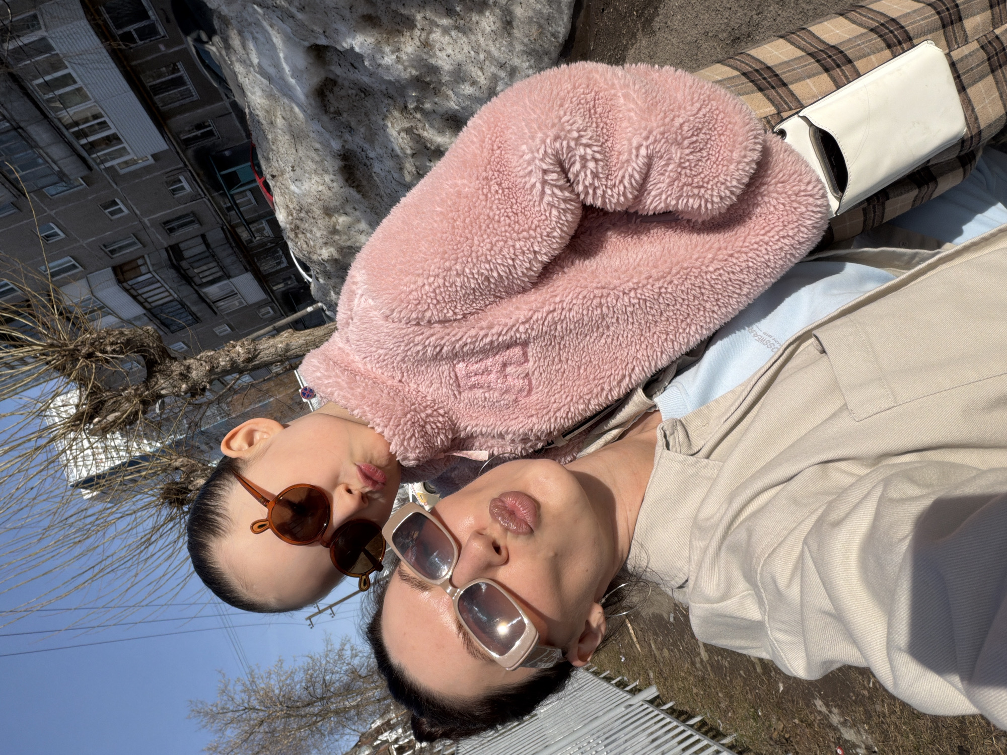 The image shows two individuals, likely a woman and a child, posing for a selfie in Perm, Russia. The woman, in the foreground, wears a beige jacket and large, square sunglasses. The child, positioned above and to the left of the woman, wears round, brown sunglasses and a pink, fuzzy jacket. Both subjects are puckering their lips, suggesting they are attempting a "duck face" pose.
In the background, a partially melted snowdrift is visible, along with bare trees and a building with multiple windows, all characteristic of a typical urban environment. A metal fence can be seen in the lower right corner. The clear blue sky indicates a sunny day.
