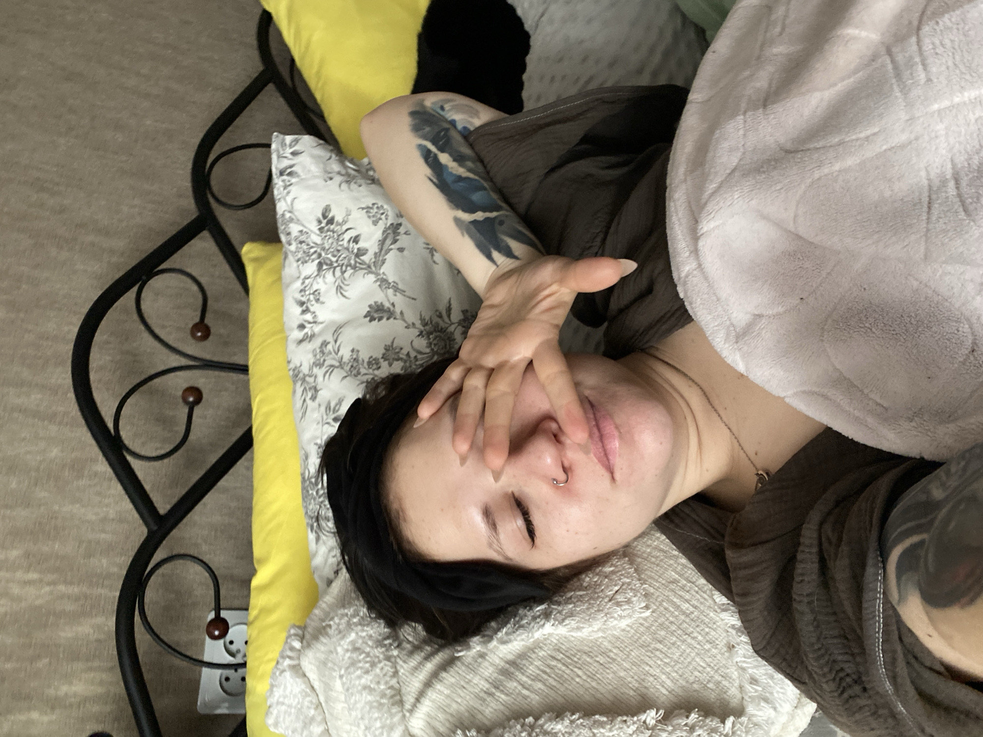 The image shows a person lying in bed. The person has dark hair, closed eyes, and a nose ring. Their hand is covering part of their face. They are lying on a white textured blanket with a floral-patterned pillow and a yellow pillow behind them. A light grey blanket is partially covering the person's body. The person has tattoos on both arms. A portion of a black wrought-iron bed frame is visible on the left, along with a wall outlet. The location is potentially in Chaikovsky, Russia.