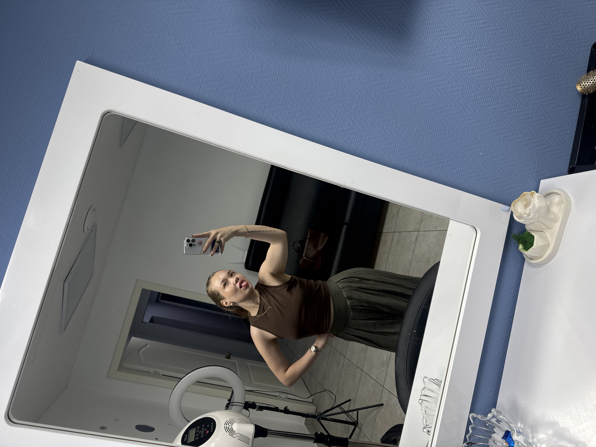 The image depicts a woman taking a selfie in a mirror. She is wearing a brown sleeveless top and an olive green skirt. The mirror reflects a room with a sofa, a door, and a ring light on a stand. On the right side of the mirror, there is a white table with a decorative item and crystal glass. The wall behind the mirror is blue. The location of the image is suggested as Chaikovsky, Russia.