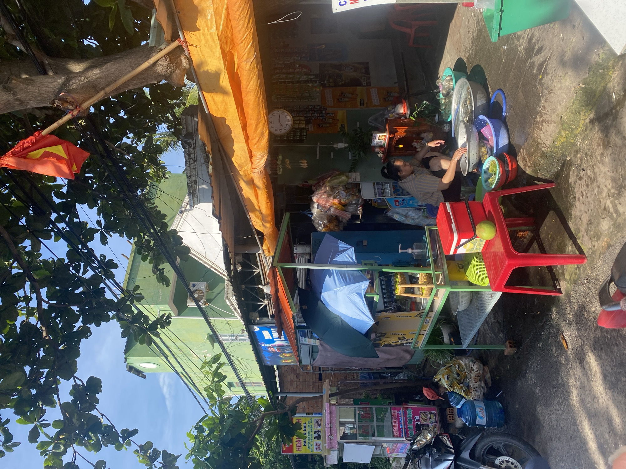 This is a street scene in Mũi Né, Vietnam, showing a small roadside stall. A person is kneeling down and appears to be washing something in basins. The stall is covered by an orange tarp and displays various items for sale, including packaged goods and what appear to be fresh produce. A motorbike is parked to the left of the stall. A green building is visible in the background. A Vietnamese flag is affixed to a pole tied to a tree. The lighting suggests it is daytime with direct sunlight.
