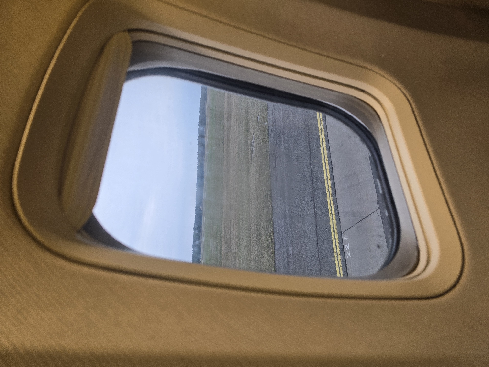 The image shows a view from an airplane window, looking out onto an airfield. The window frame is a light tan color and has a textured surface. Outside the window, there is a tarmac with yellow painted lines, and beyond that, a large grassy field. In the distance, a line of trees forms the horizon. The sky is overcast. The airfield appears to be part of Warsaw Modlin Airport (WMI), located near Nowy Dwór Mazowiecki, Poland.