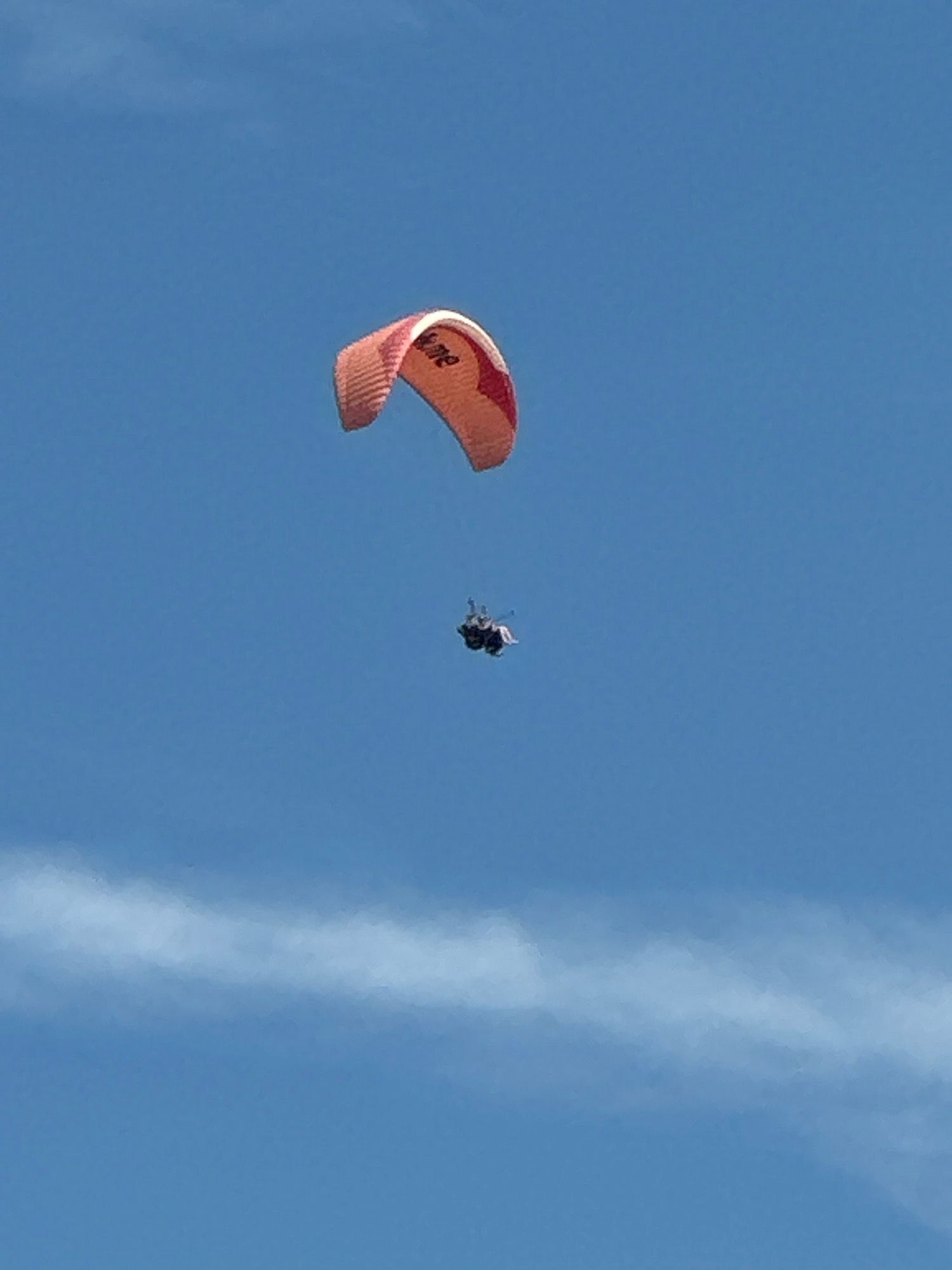 A paraglider with the word "ZONE" visible on its wing soars through a clear blue sky. Beneath the paraglider, two individuals are suspended in a harness. A faint white cloud streak stretches horizontally across the lower portion of the frame. The scene appears to be an aerial activity occurring in or near Boreti, Montenegro.